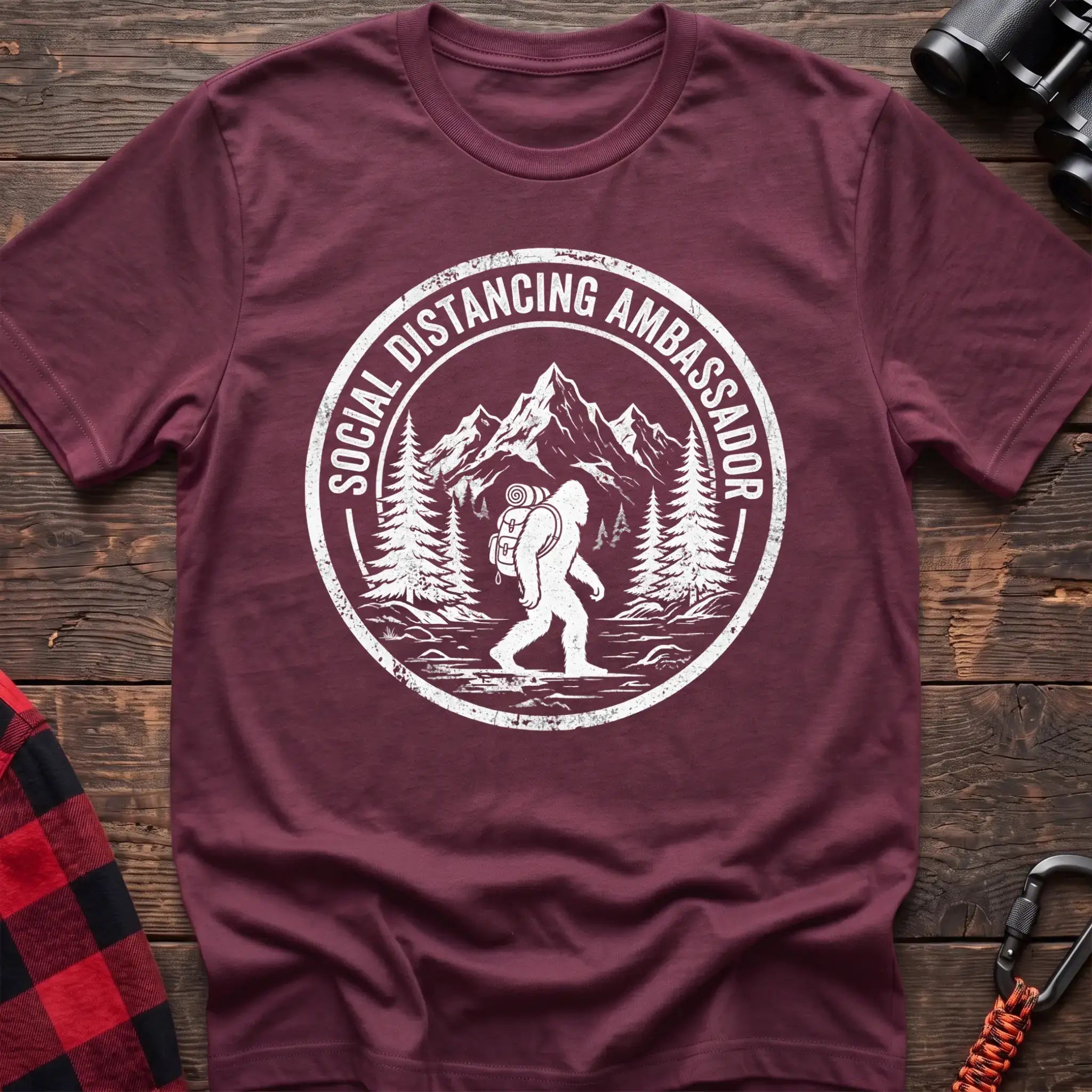 Heather maroon t-shirt with white circular badge graphic of Bigfoot hiking with a backpack through mountains and the text Social Distancing Ambassador