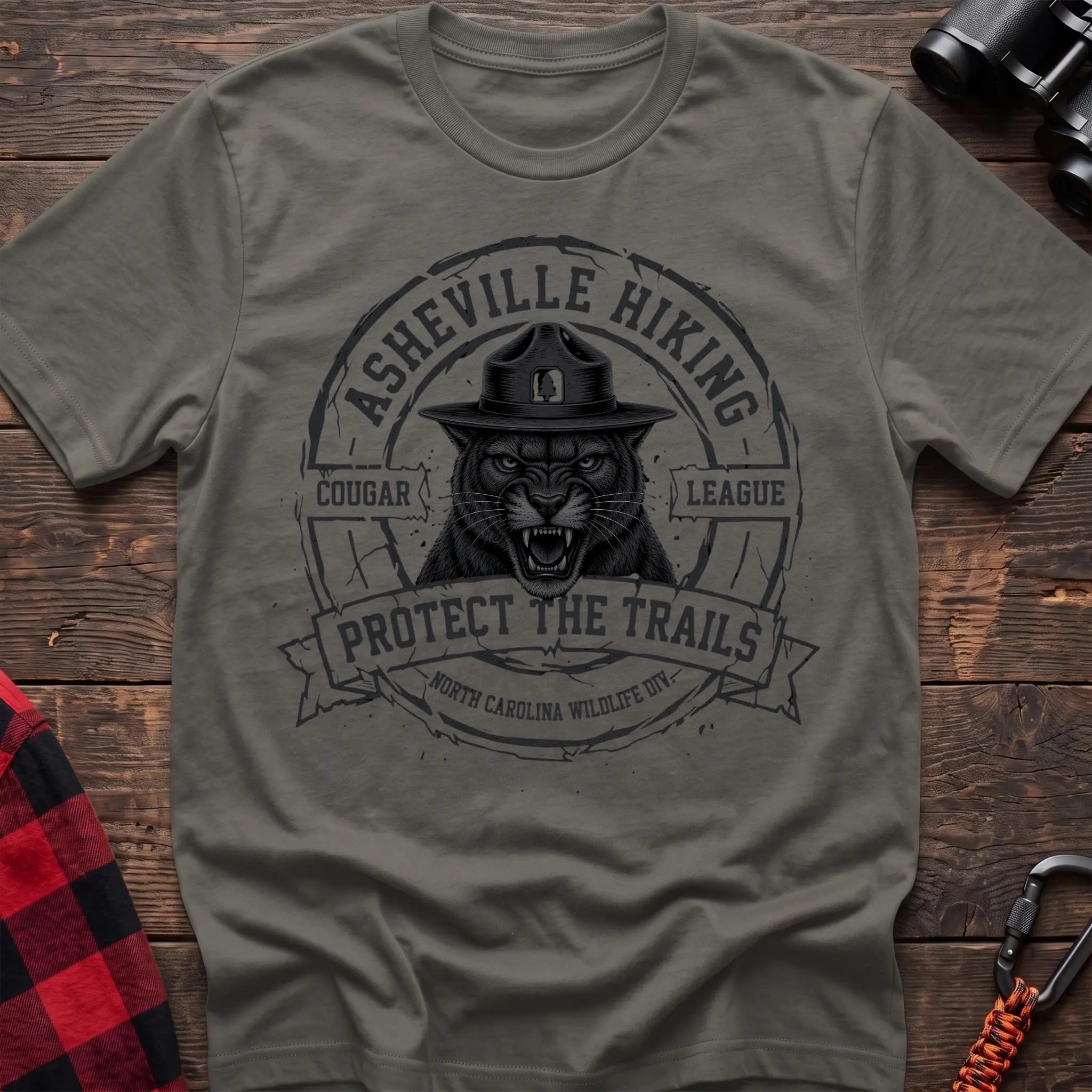 Heather military green cotton t-shirt with vintage Asheville Hiking Cougar League graphic of a roaring cougar in a park ranger hat, Protect the Trails North Carolina Wildlife Division badge, flat lay on rustic wood with hiking gear