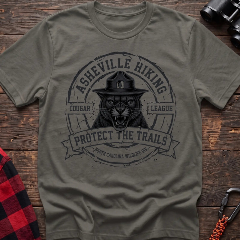 Heather military green cotton t-shirt with vintage Asheville Hiking Cougar League graphic of a roaring cougar in a park ranger hat, Protect the Trails North Carolina Wildlife Division badge, flat lay on rustic wood with hiking gear