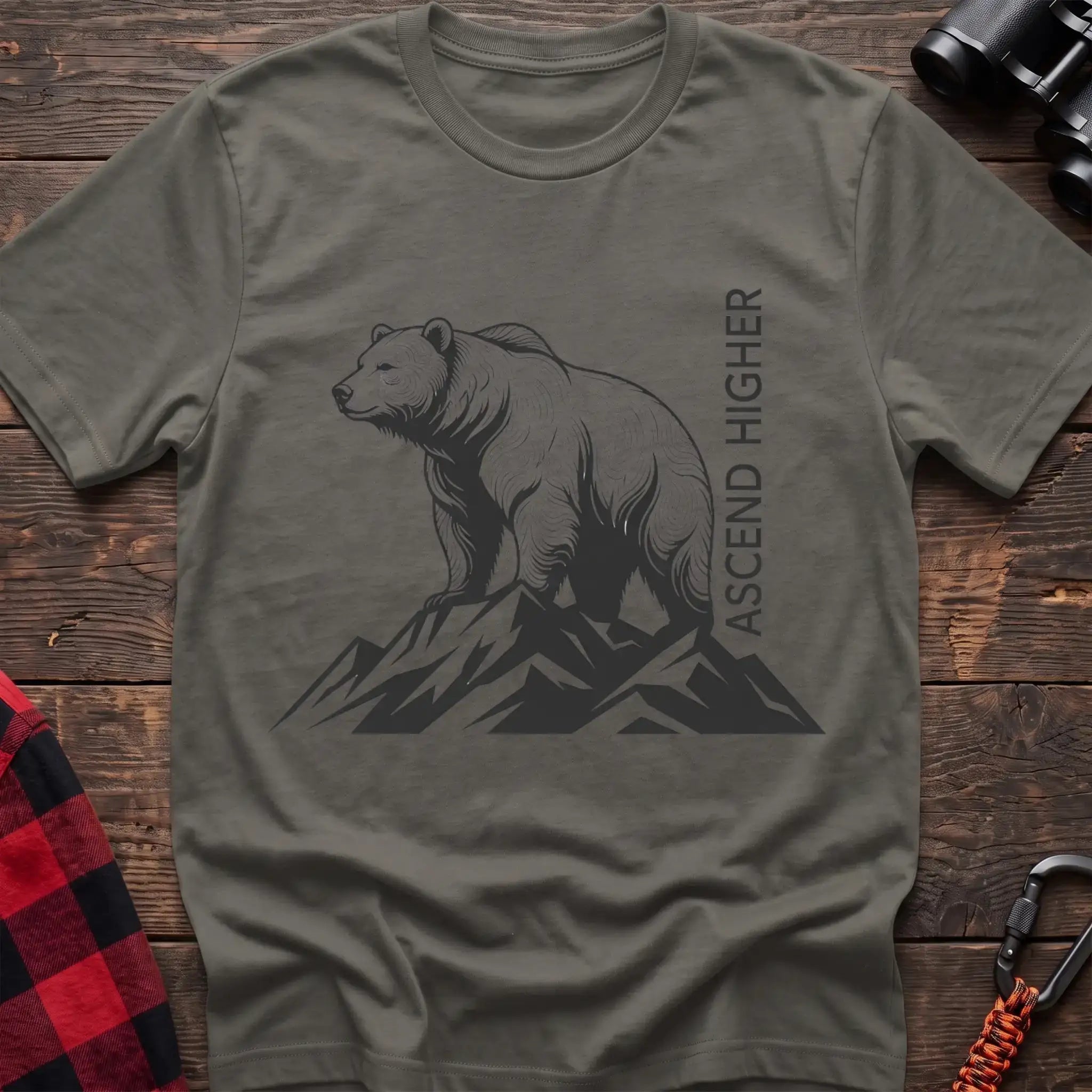 Heather military green t-shirt with a black illustration of a bear standing on mountain peaks, with the vertical text “ASCEND HIGHER” beside it, laid flat on a wooden surface.