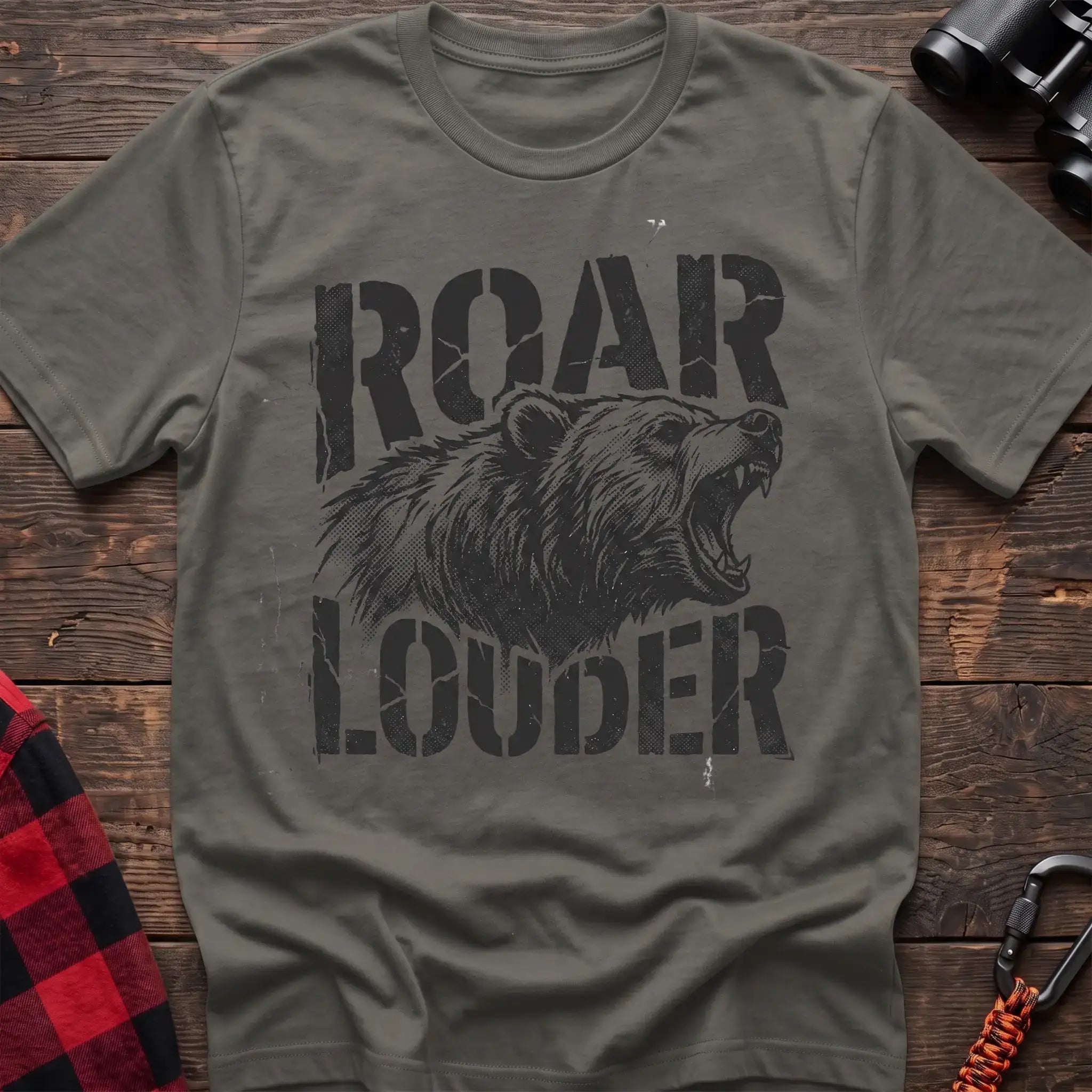 Heather military green t-shirt with a distressed black graphic of a roaring bear head between the bold text “ROAR” and “LOUDER,” laid flat on a wooden surface.