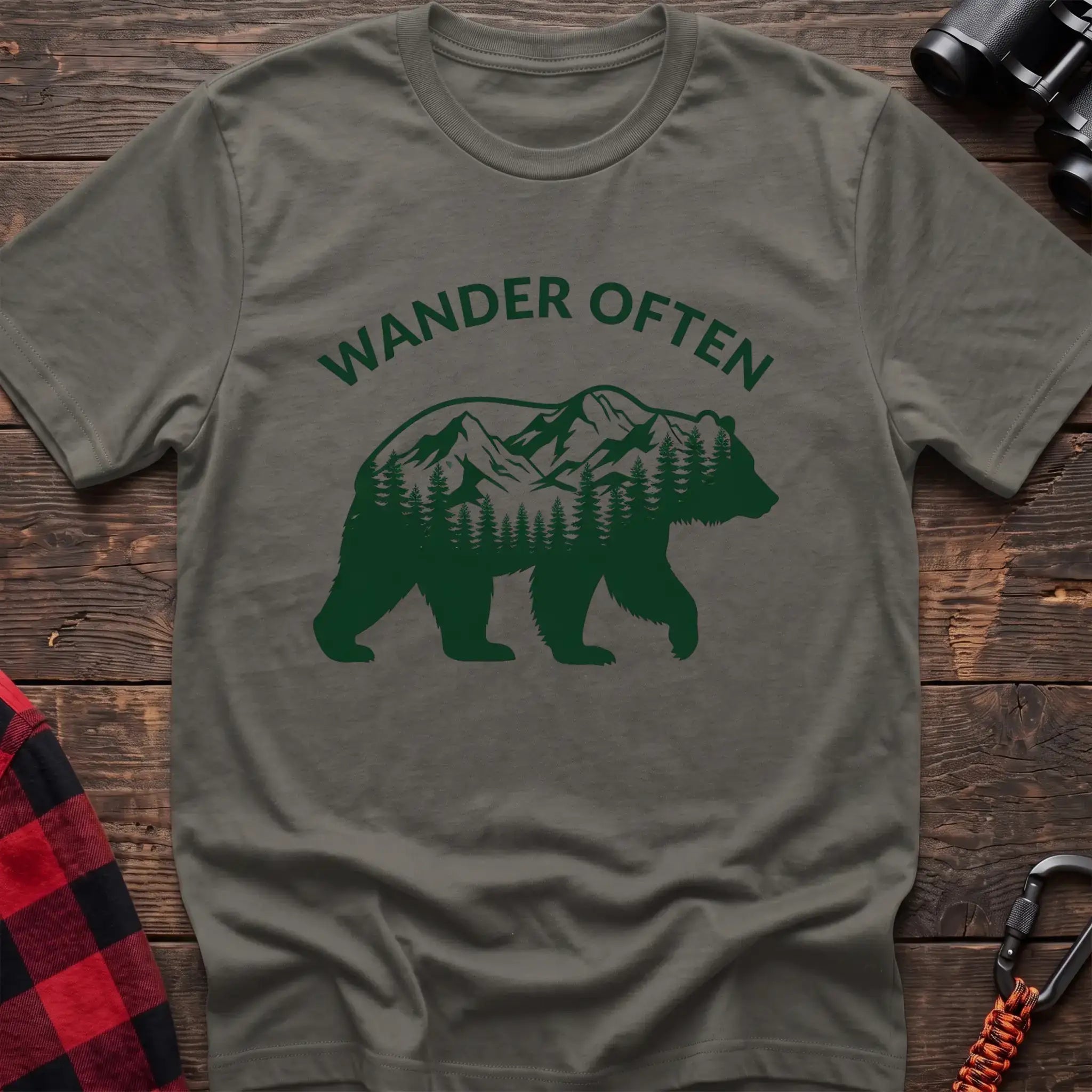 Heather military green t-shirt with a dark green silhouette of a walking bear containing a mountain and pine forest scene inside, with the arched text “WANDER OFTEN” above it, laid flat on a wooden surface.