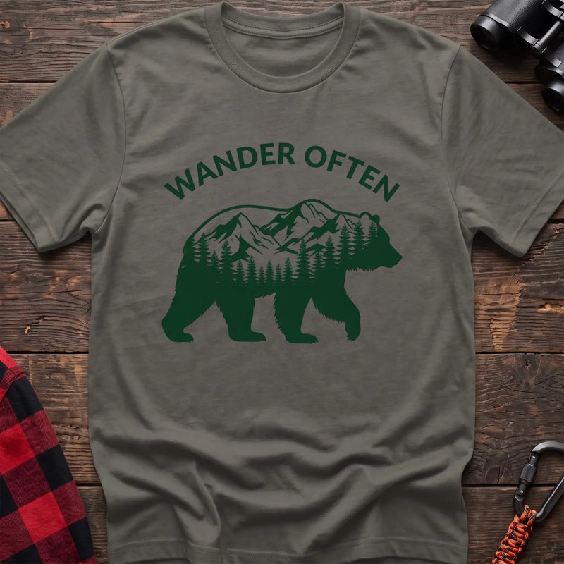 Heather military green t-shirt with a dark green silhouette of a walking bear containing a mountain and pine forest scene inside, with the arched text “WANDER OFTEN” above it, laid flat on a wooden surface.