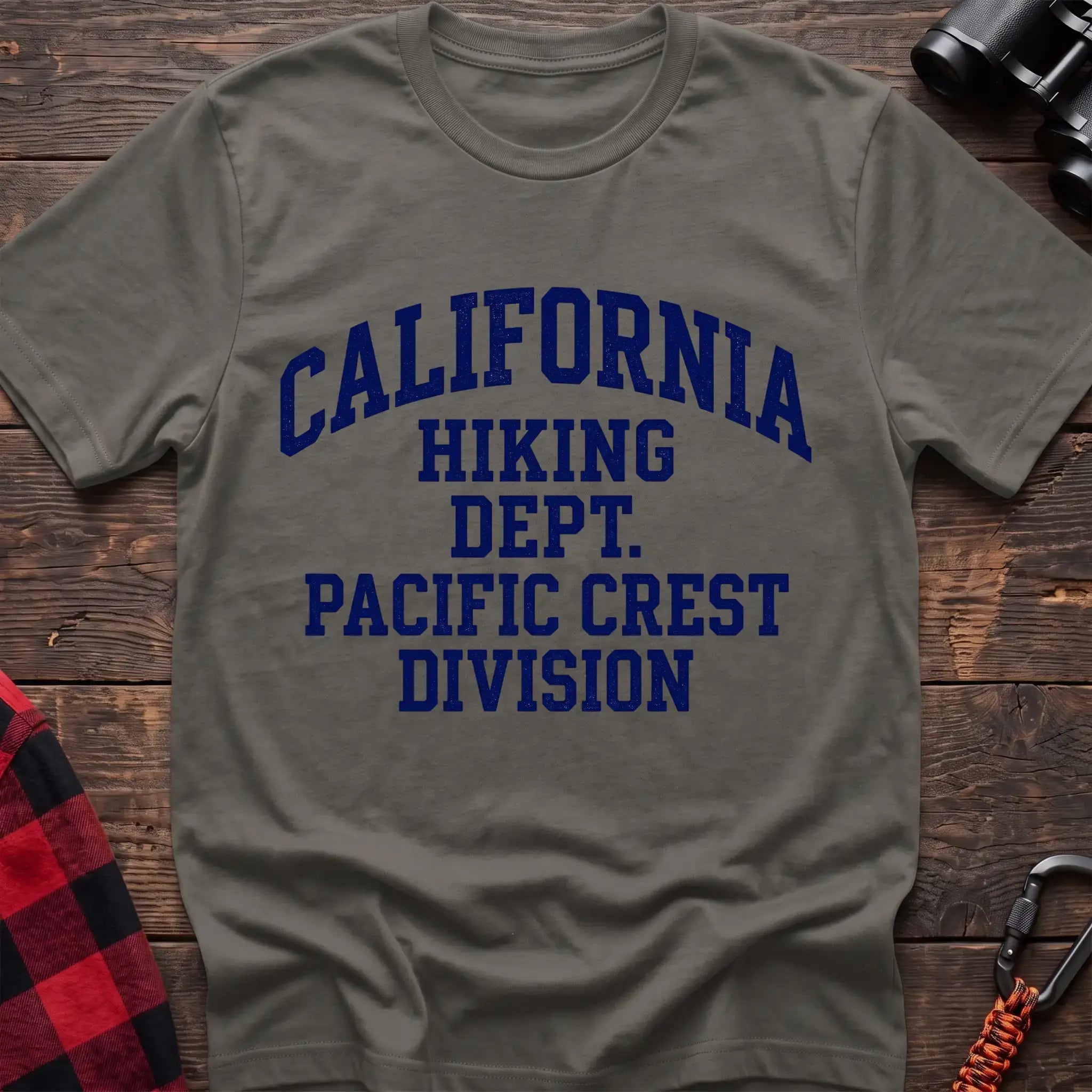 Heather military green t-shirt with distressed blue collegiate-style text reading “CALIFORNIA HIKING DEPT. PACIFIC CREST DIVISION,” laid flat on a wooden surface.