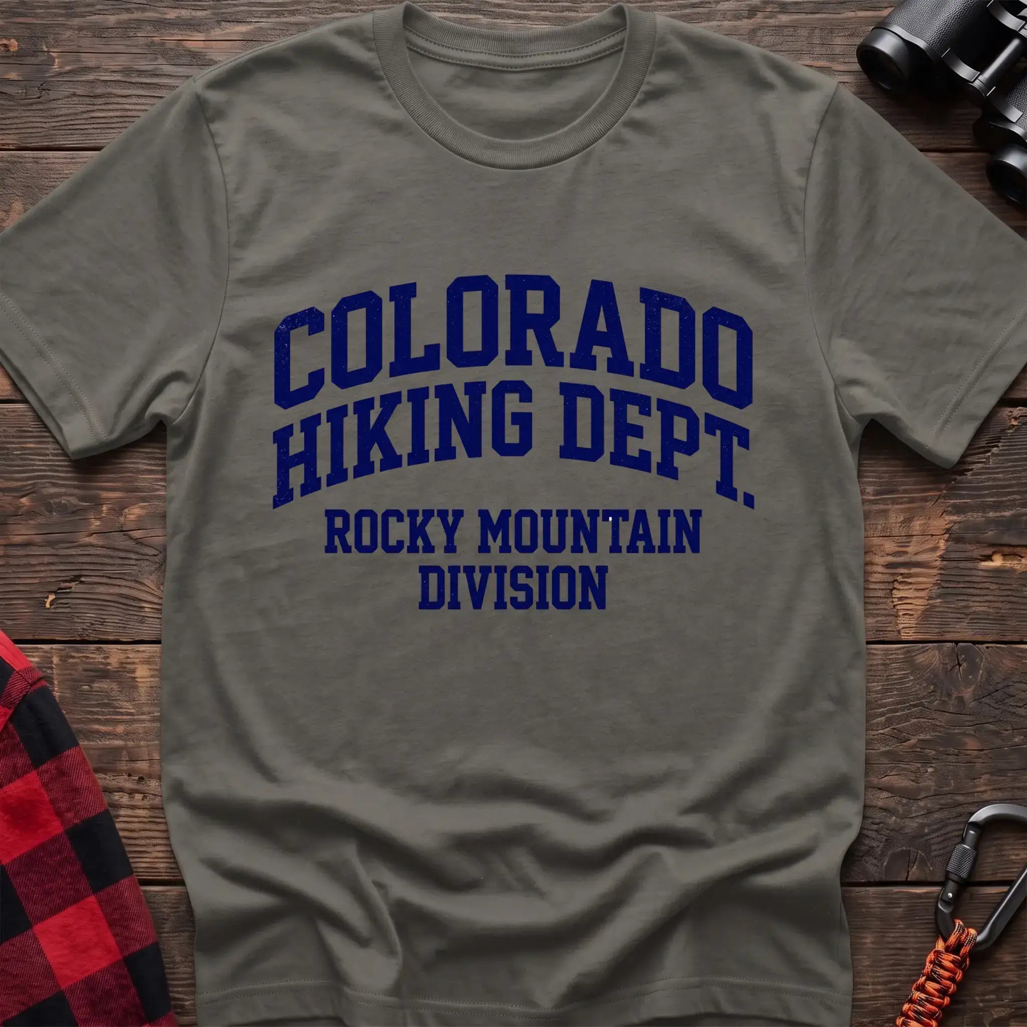 Heather military green t-shirt with distressed dark blue collegiate-style text reading “COLORADO HIKING DEPT. ROCKY MOUNTAIN DIVISION,” laid flat on a wooden surface.