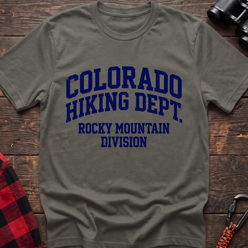Heather military green t-shirt with distressed dark blue collegiate-style text reading “COLORADO HIKING DEPT. ROCKY MOUNTAIN DIVISION,” laid flat on a wooden surface.