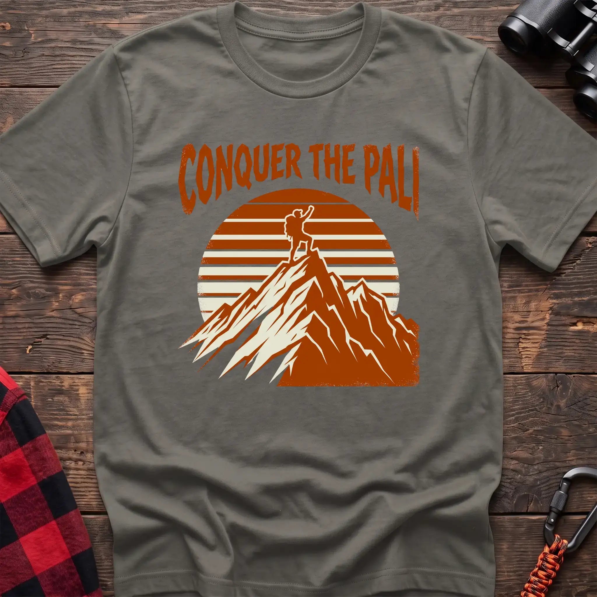 Heather military green t-shirt with orange text reading Conquer the Pali above a hiker standing on a mountain peak with a retro sun graphic