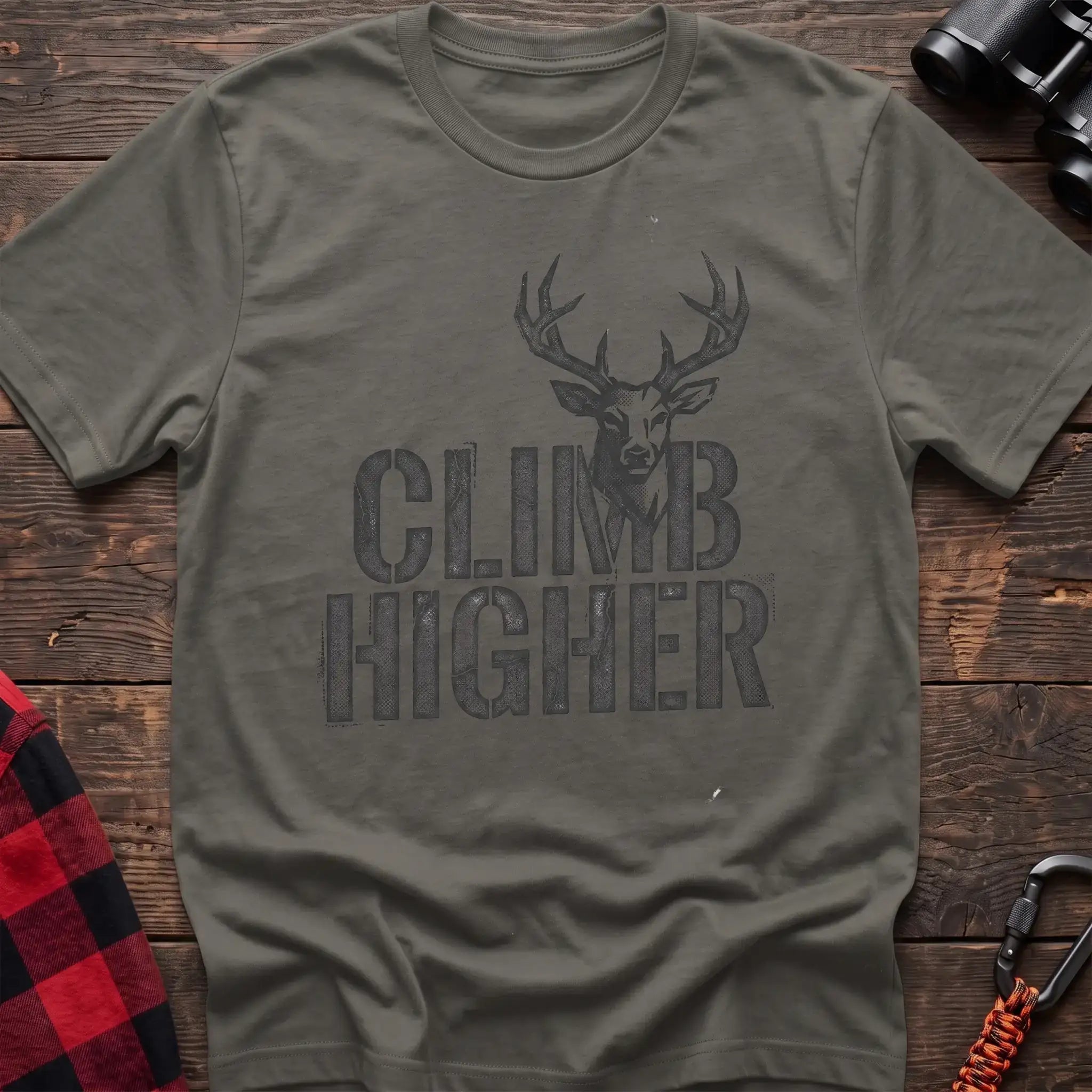 Heather military green t-shirt with a distressed dark graphic of a deer’s head and antlers rising from the bold text “CLIMB HIGHER,” laid flat on a wooden surface.