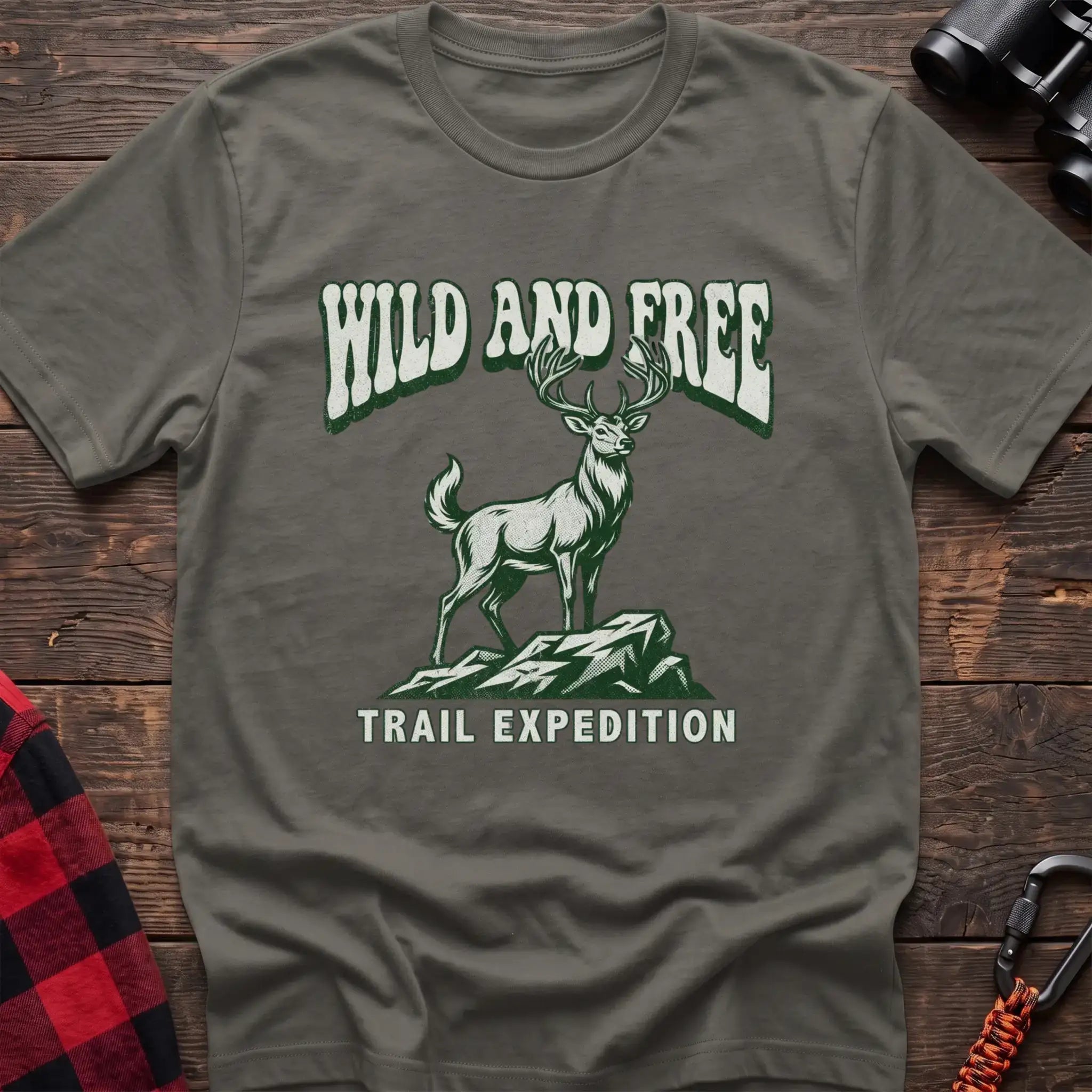 Heather military green t-shirt with a rustic graphic of a deer standing on a rocky outcrop, with the arched text “WILD AND FREE” above and “TRAIL EXPEDITION” below, laid flat on a wooden surface.
