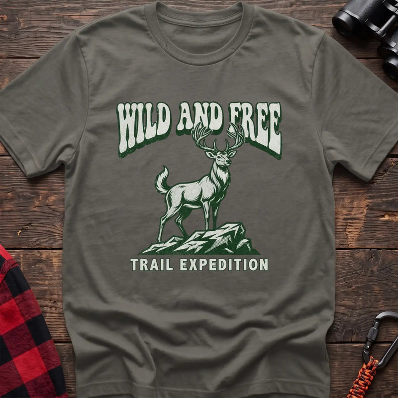 Heather military green t-shirt with a rustic graphic of a deer standing on a rocky outcrop, with the arched text “WILD AND FREE” above and “TRAIL EXPEDITION” below, laid flat on a wooden surface.