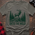 Heather military green t-shirt with a dark green illustration of an elk standing on a rocky outcrop among pine trees, with the text “INTO THE WILD” printed below, laid flat on a wooden surface.