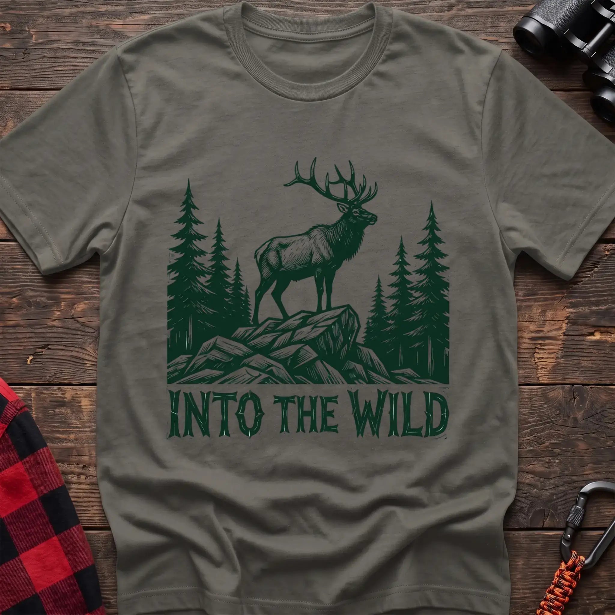 Heather military green t-shirt with a dark green illustration of an elk standing on a rocky outcrop among pine trees, with the text “INTO THE WILD” printed below, laid flat on a wooden surface.