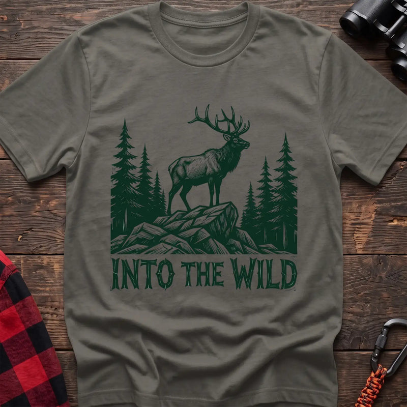 Heather military green t-shirt with a dark green illustration of an elk standing on a rocky outcrop among pine trees, with the text “INTO THE WILD” printed below, laid flat on a wooden surface.