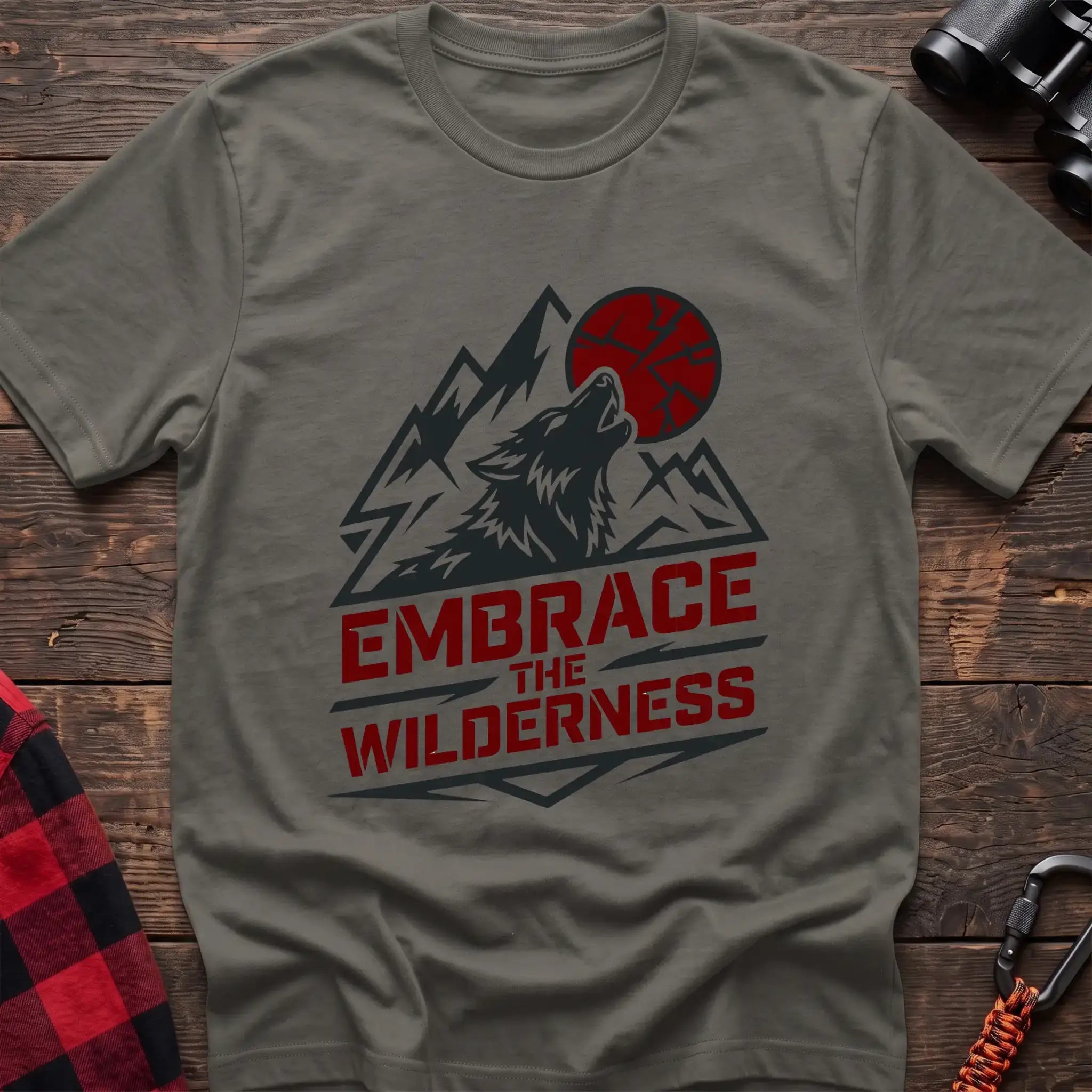 Heather military green t-shirt with a dark graphic of a howling wolf against mountain peaks and a red sun, with the bold red text “EMBRACE THE WILDERNESS” below, laid flat on a wooden surface.