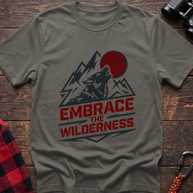 Heather military green t-shirt with a dark graphic of a howling wolf against mountain peaks and a red sun, with the bold red text “EMBRACE THE WILDERNESS” below, laid flat on a wooden surface.