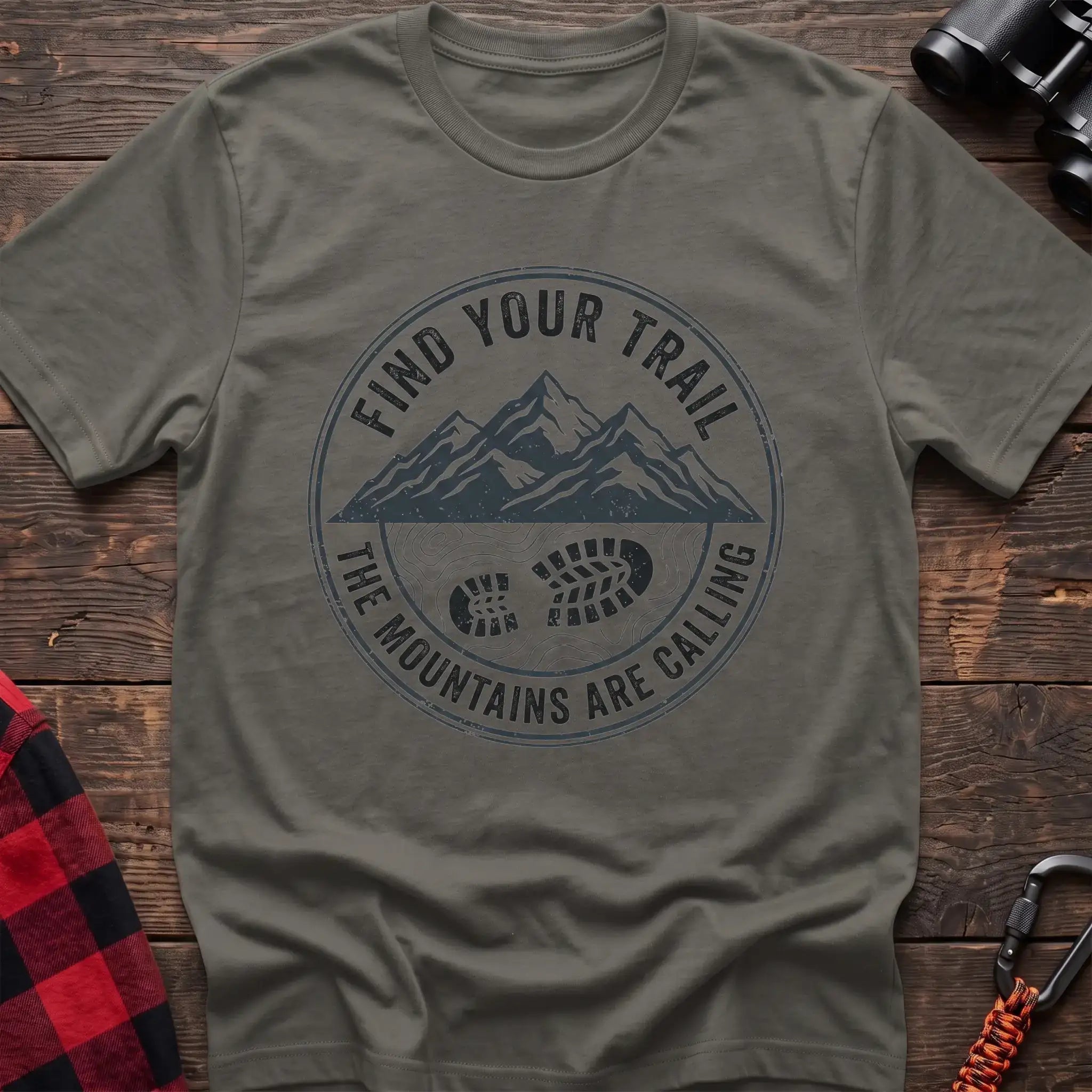 Heather military green t-shirt with a circular dark gray badge design featuring mountain peaks above a hiking boot print on topographic lines, with the text “FIND YOUR TRAIL” and “THE MOUNTAINS ARE CALLING,” laid flat on a wooden surface.