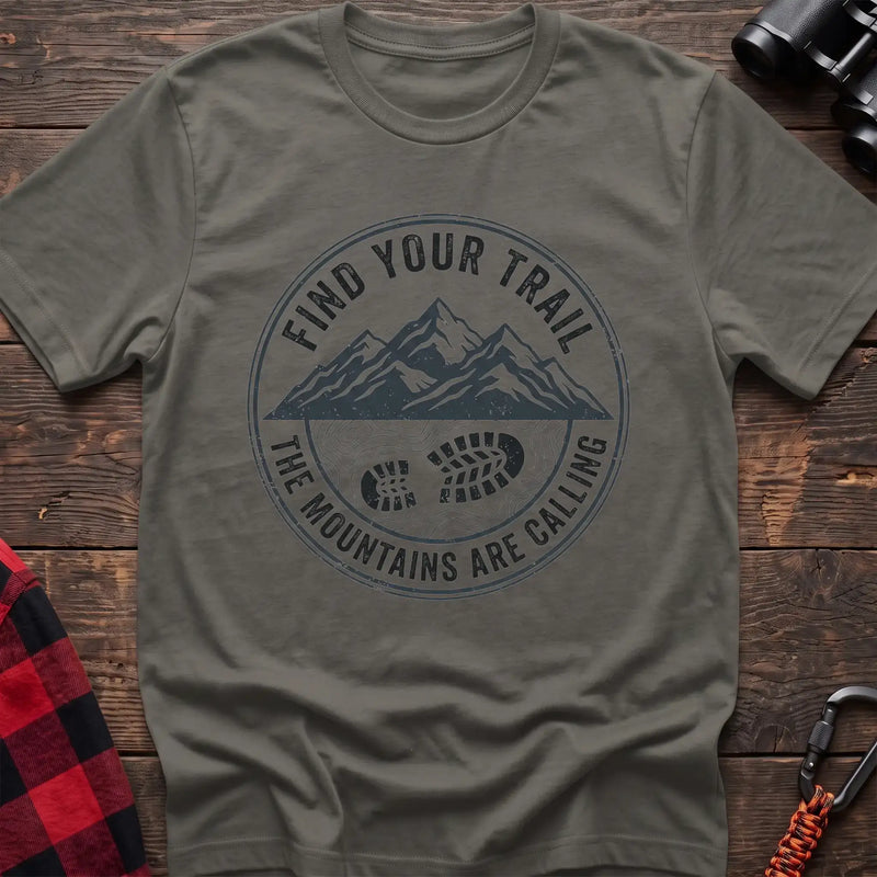 Heather military green t-shirt with a circular dark gray badge design featuring mountain peaks above a hiking boot print on topographic lines, with the text “FIND YOUR TRAIL” and “THE MOUNTAINS ARE CALLING,” laid flat on a wooden surface.
