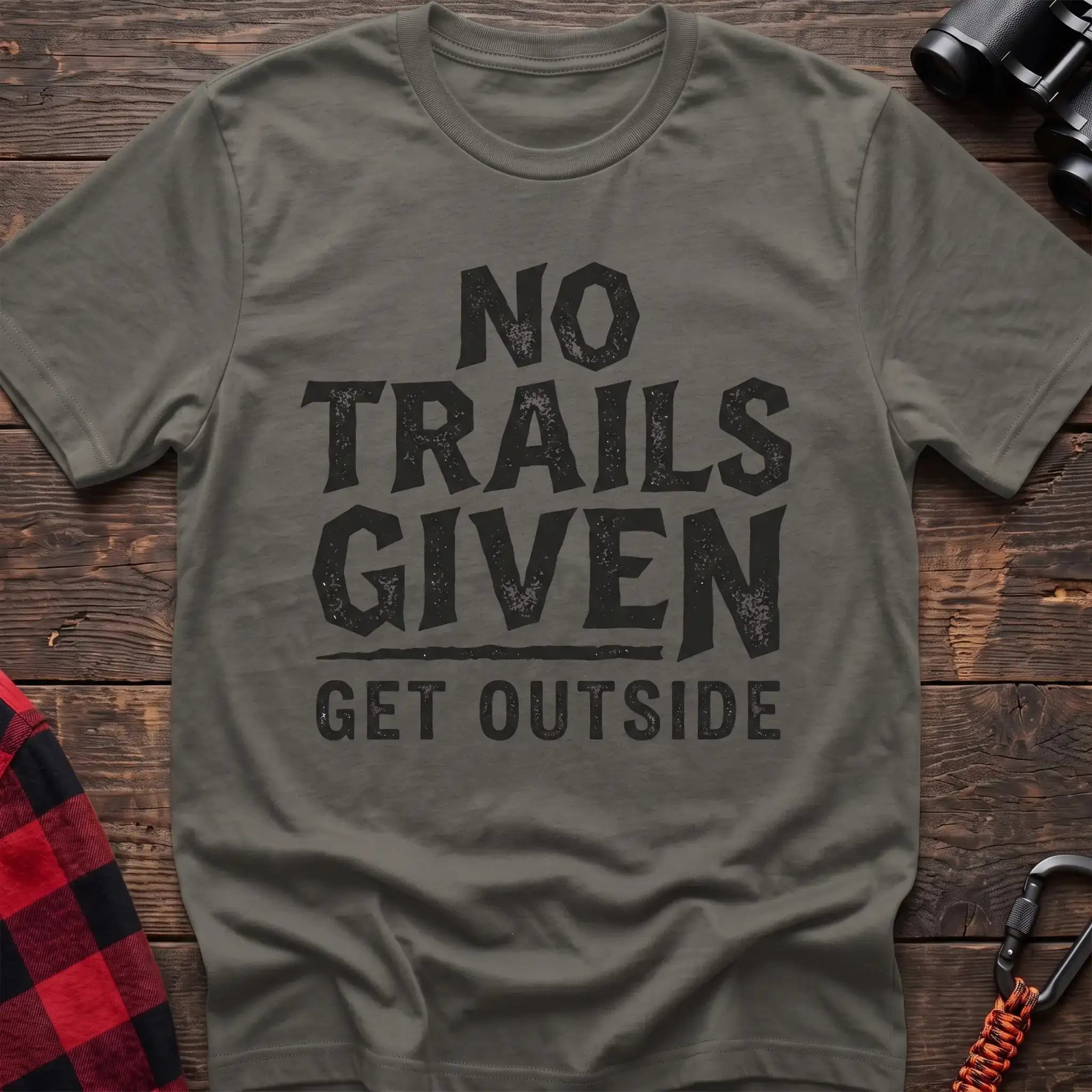Heather military green t-shirt with distressed black text reading “NO TRAILS GIVEN” and “GET OUTSIDE” below it, laid flat on a wooden surface.