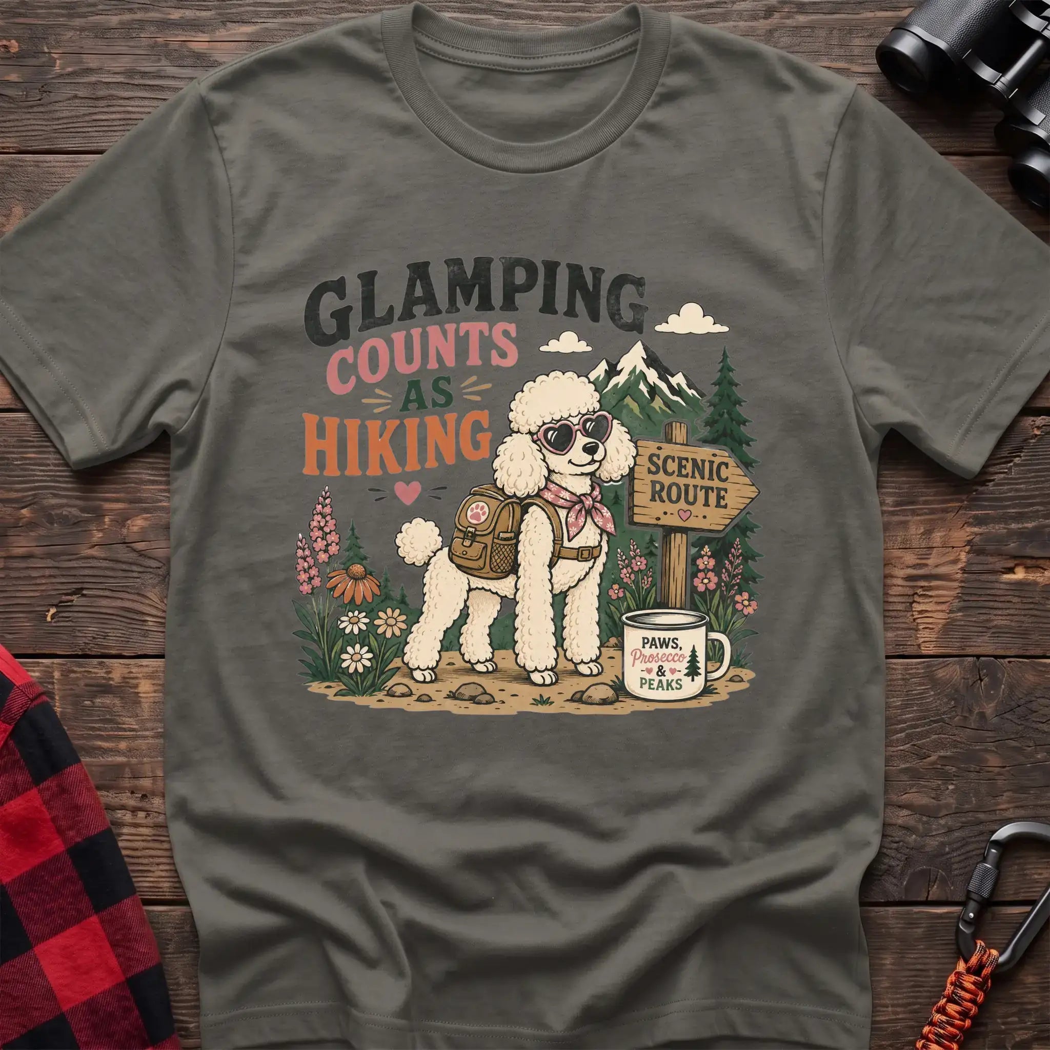 Heather military green t-shirt with a colorful illustration of a poodle in sunglasses and a backpack on a scenic trail, with the text “GLAMPING COUNTS AS HIKING,” laid flat on a wooden surface.
