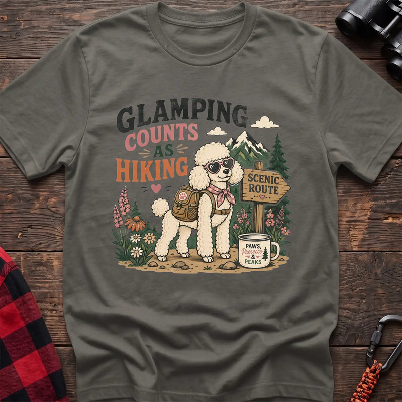 Heather military green t-shirt with a colorful illustration of a poodle in sunglasses and a backpack on a scenic trail, with the text “GLAMPING COUNTS AS HIKING,” laid flat on a wooden surface.