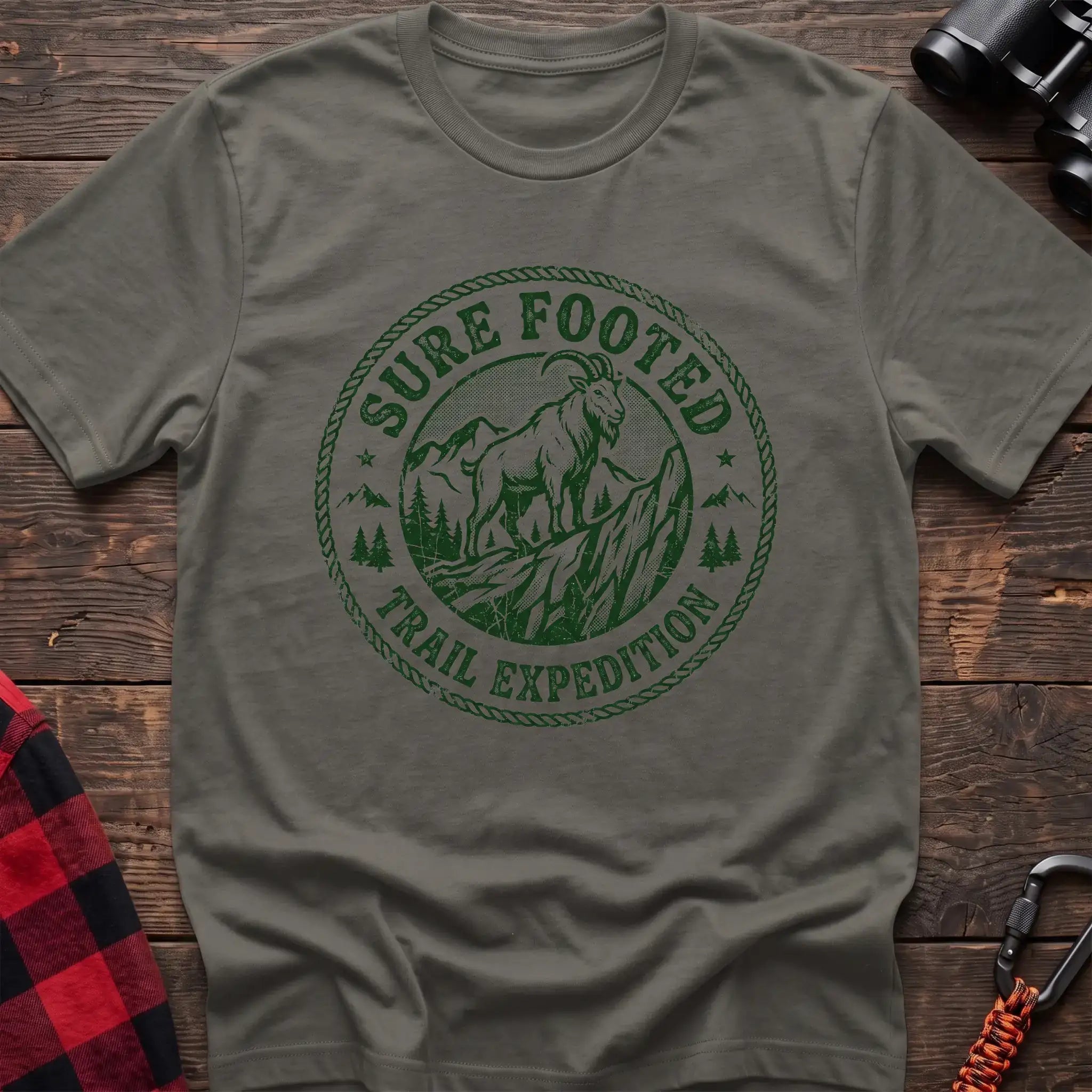 Heather military green t-shirt with circular badge graphic of a mountain goat on a rocky peak reading Sure Footed Trail Expedition