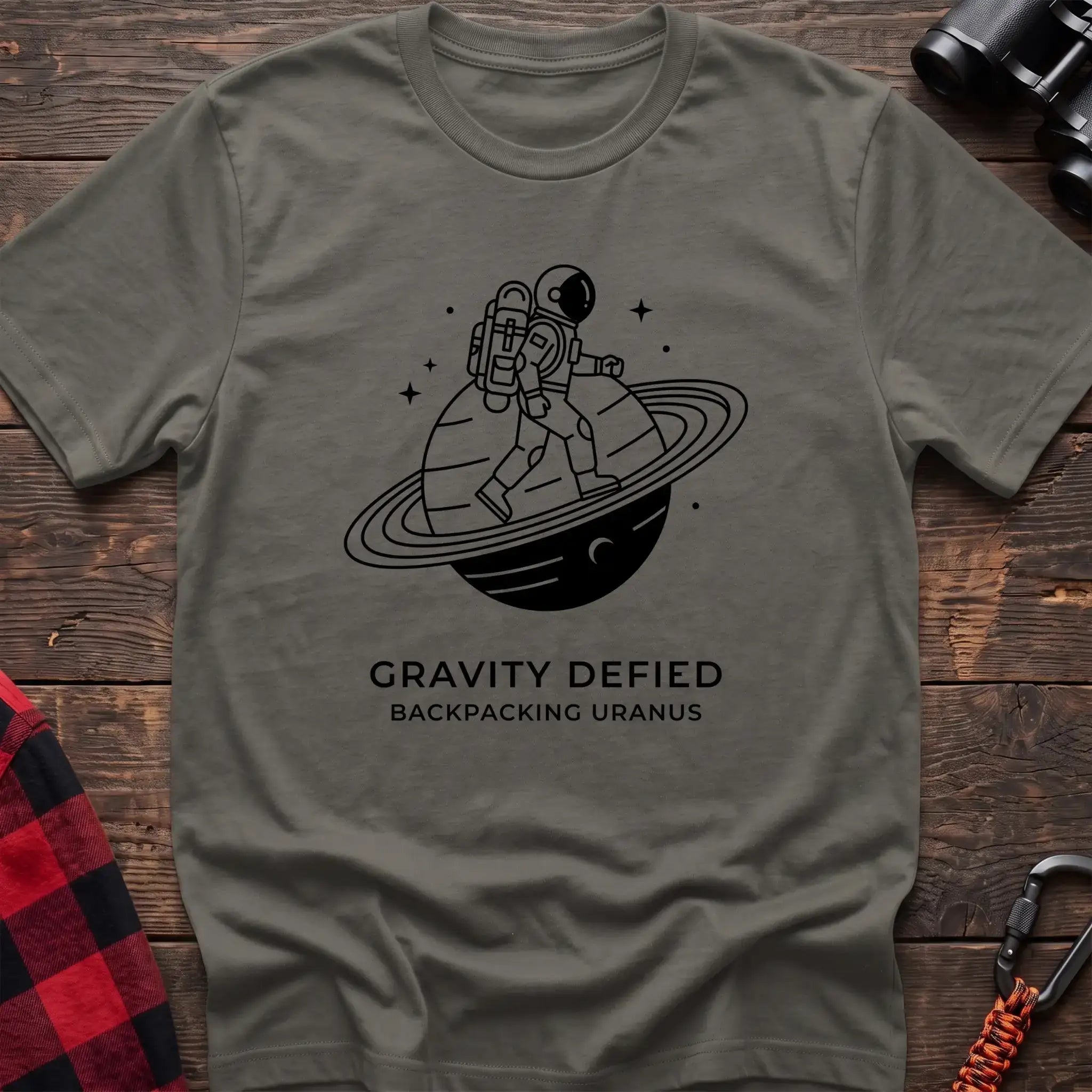 Heather military green crew-neck t-shirt with black line-art graphic of an astronaut backpacking across a ringed planet surrounded by stars, with the text “GRAVITY DEFIED / BACKPACKING URANUS” printed below, laid flat on a wooden surface.