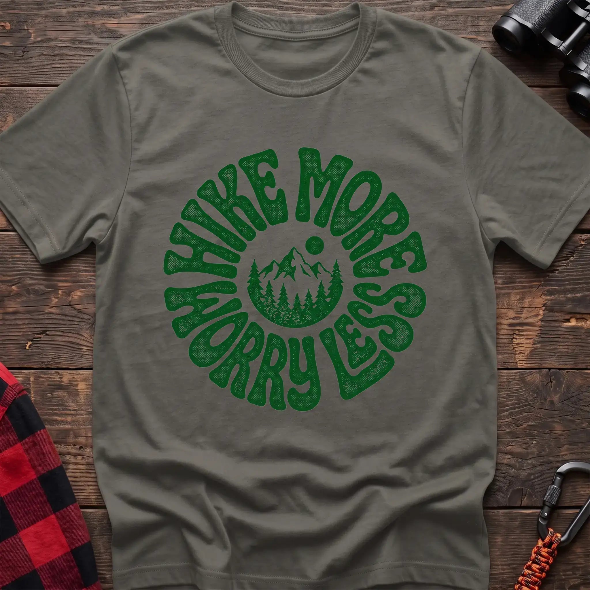 Heather military green t-shirt with a circular green design featuring mountains and pine trees in the center, surrounded by the text “HIKE MORE WORRY LESS,” laid flat on a wooden surface.