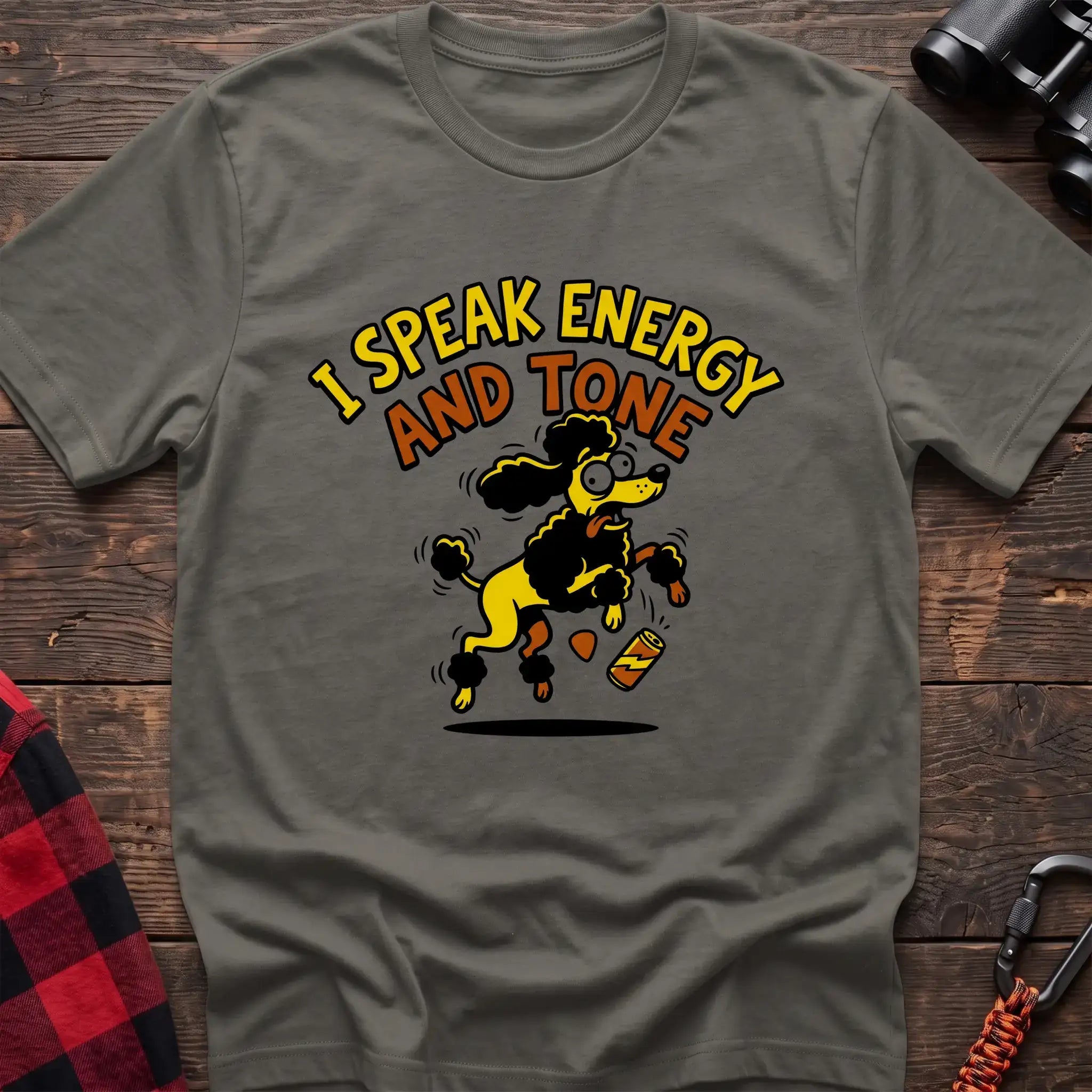 Heather military green t-shirt with a cartoon yellow and black poodle jumping excitedly next to a falling energy drink can, with the arched text “I SPEAK ENERGY AND TONE,” laid flat on a wooden surface.