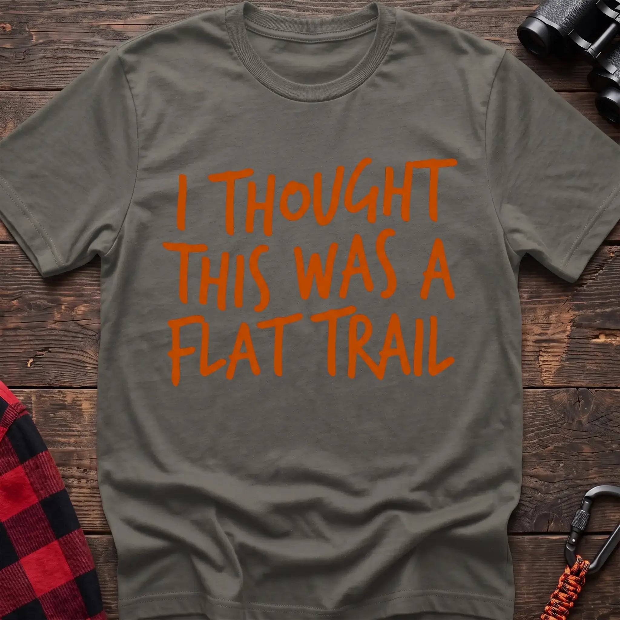Heather military green t-shirt with bold orange handwritten-style text reading “I THOUGHT THIS WAS A FLAT TRAIL,” laid flat on a wooden surface.