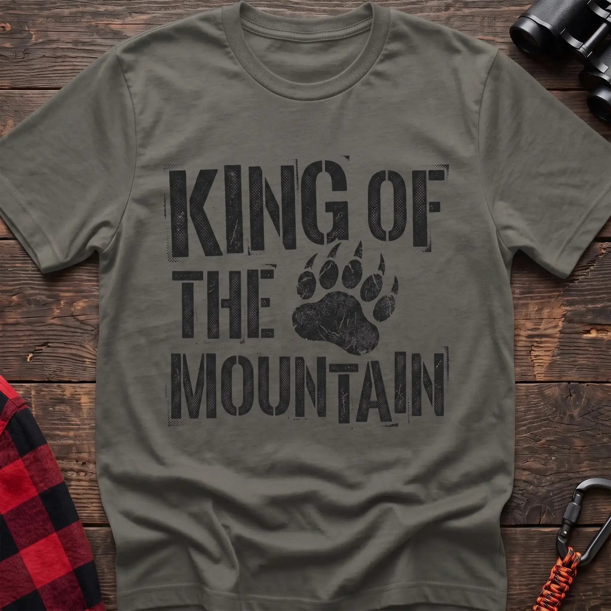 Heather military green t-shirt with distressed black text reading “KING OF THE MOUNTAIN” and a large bear paw print integrated into the design, laid flat on a wooden surface.