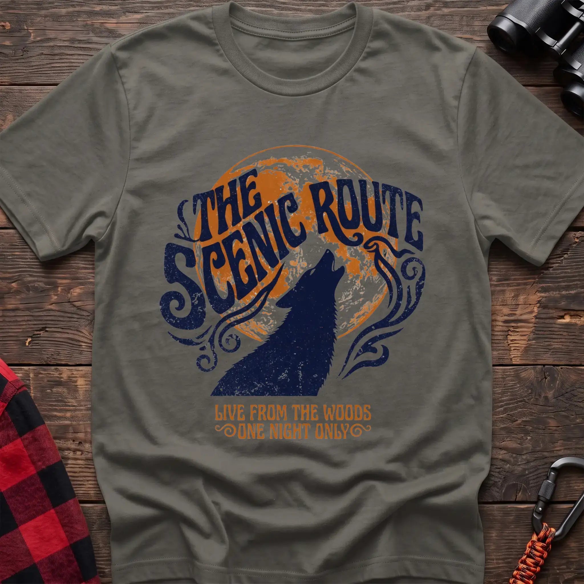 Heather military green t-shirt with a dark blue howling wolf silhouette against an orange circle, with the arched text “THE SCENIC ROUTE” above and “LIVE FROM THE WOODS / ONE NIGHT ONLY” below, laid flat on a wooden surface.