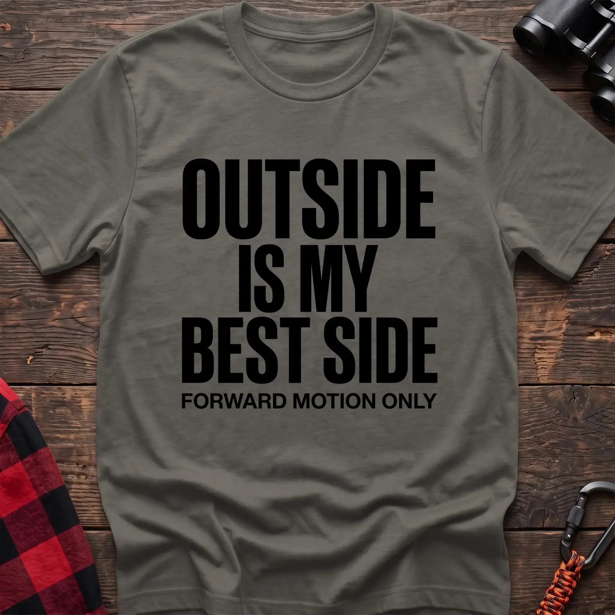 Heather military green t-shirt with bold black text reading “OUTSIDE IS MY BEST SIDE” and “FORWARD MOTION ONLY” below it, laid flat on a wooden surface.
