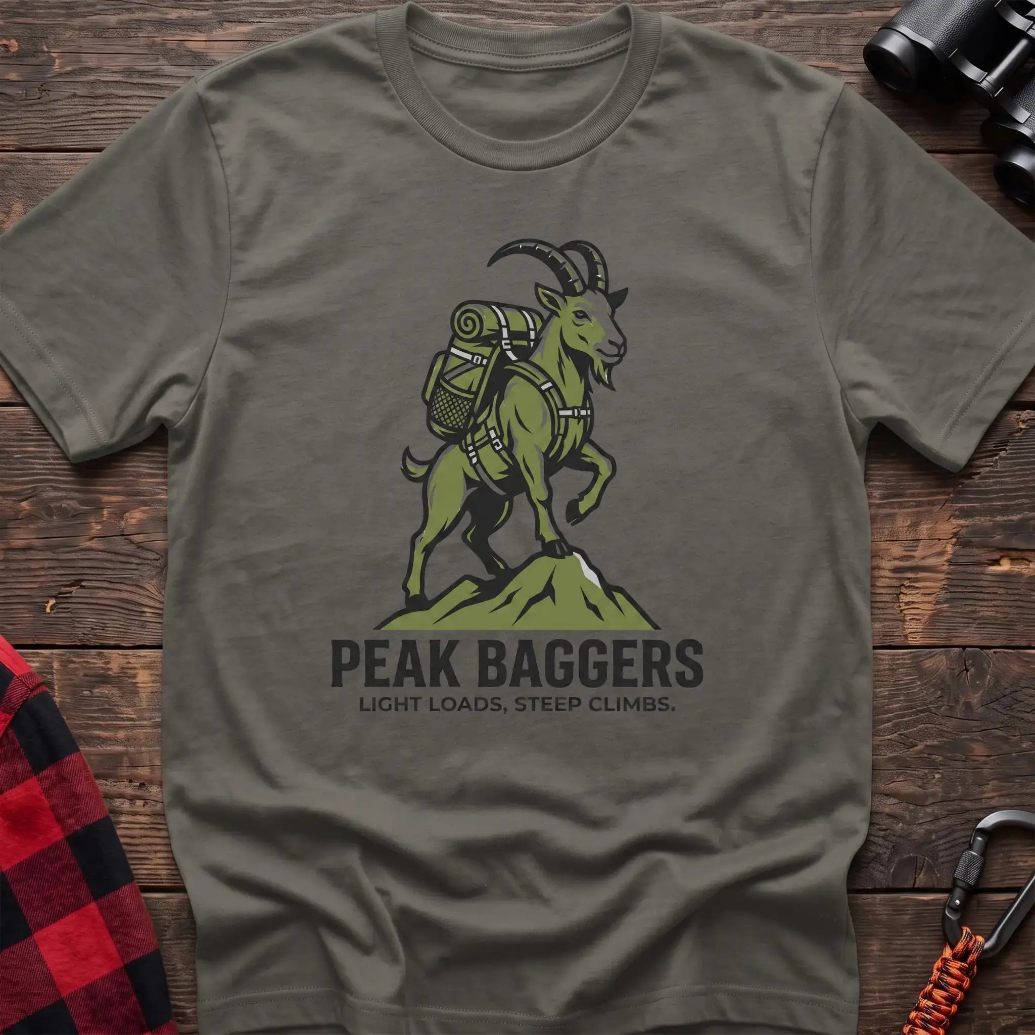 Heather military green t-shirt with an illustration of a mountain goat wearing a backpack standing on a peak, with the text “PEAK BAGGERS” and “LIGHT LOADS, STEEP CLIMBS,” laid flat on a wooden surface.