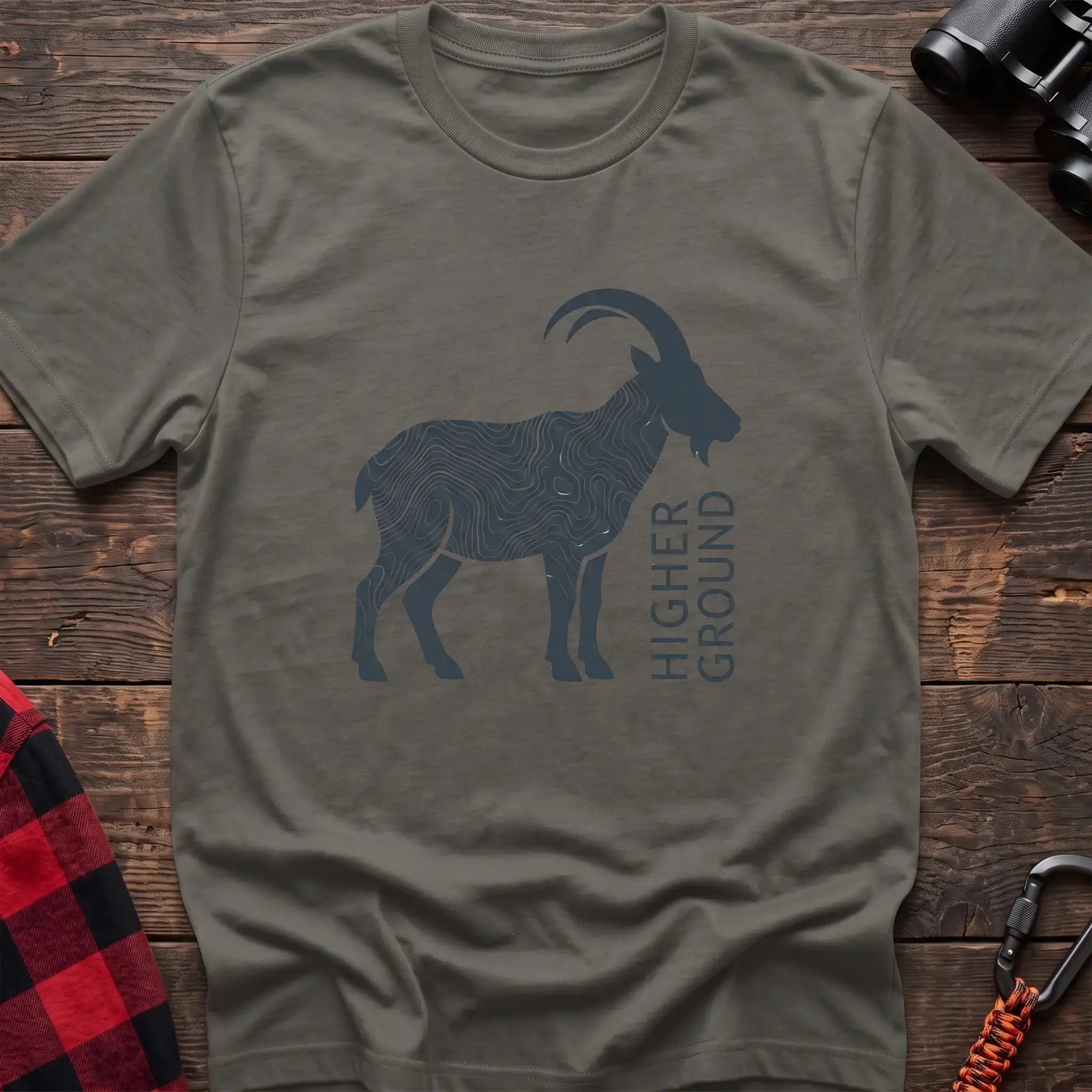 Heather military green t-shirt with a dark silhouette of a mountain goat filled with topographic contour lines, and the vertical text “HIGHER GROUND” printed beside it, laid flat on a wooden surface.