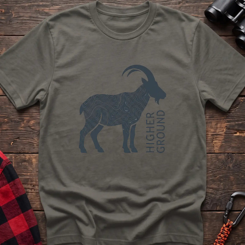 Heather military green t-shirt with a dark silhouette of a mountain goat filled with topographic contour lines, and the vertical text “HIGHER GROUND” printed beside it, laid flat on a wooden surface.