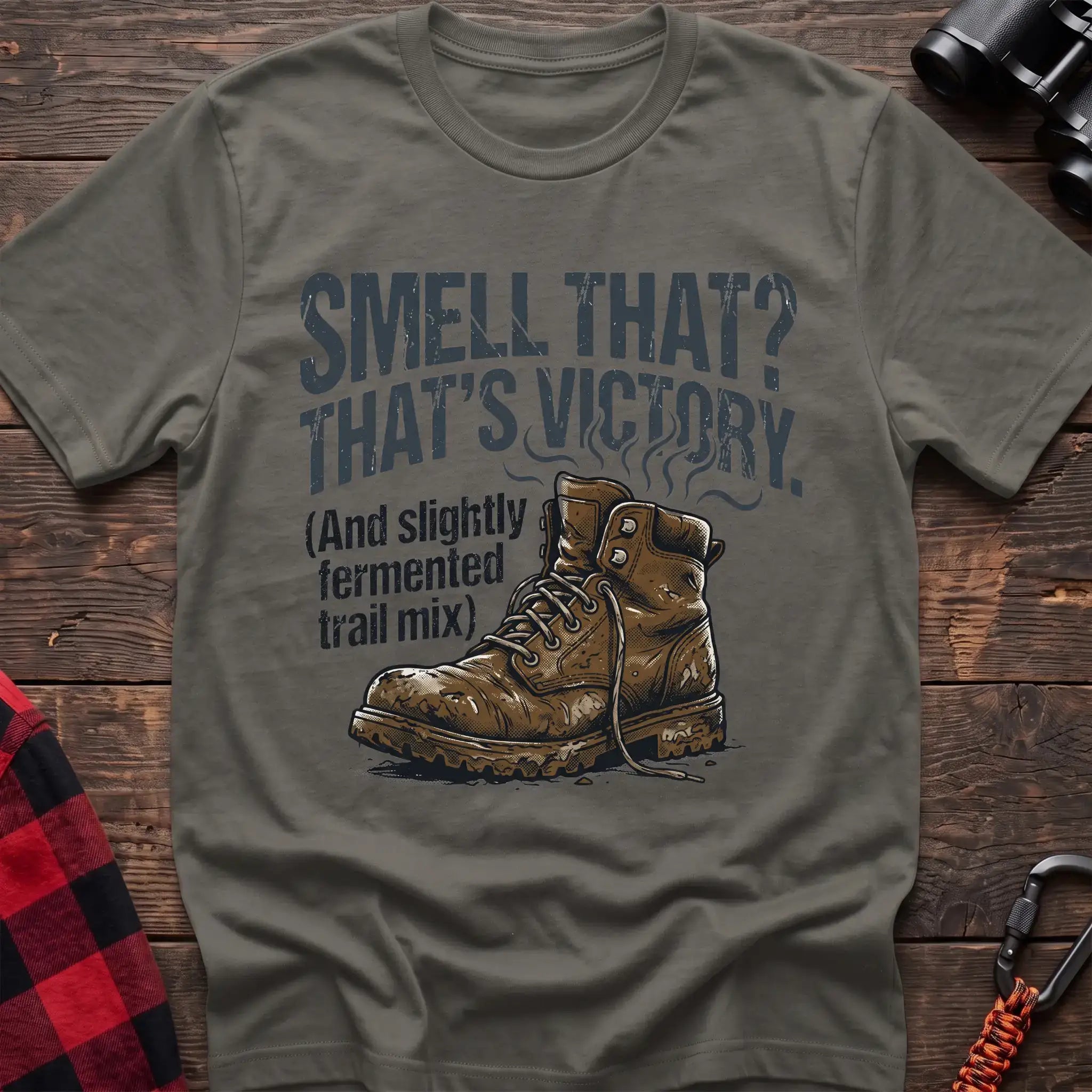 Heather military green t-shirt with an illustration of a dirty hiking boot with rising steam lines, and the text “SMELL THAT? THAT’S VICTORY. (And slightly fermented trail mix),” laid flat on a wooden surface