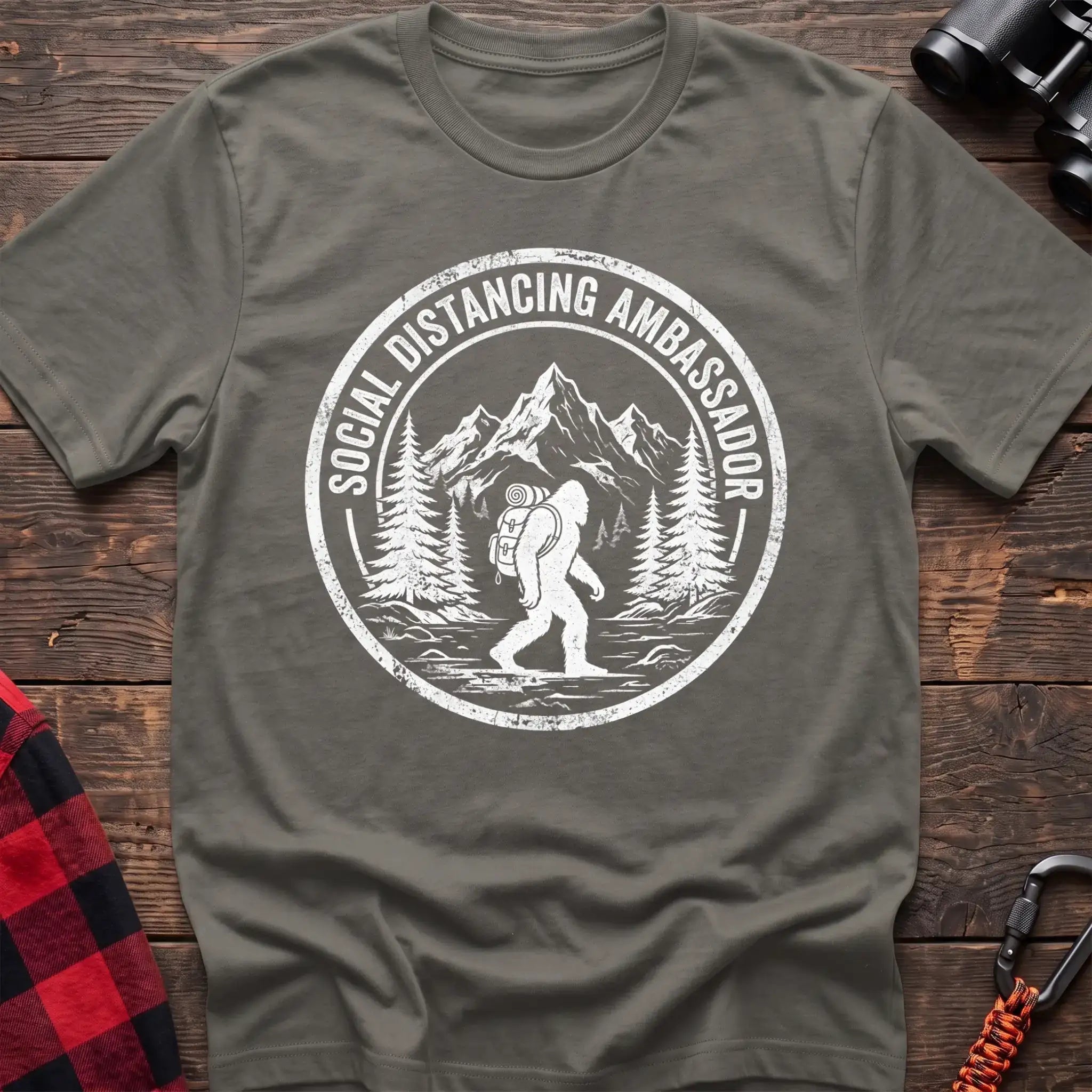 Heather military green t-shirt with white circular badge graphic of Bigfoot hiking with a backpack through mountains and the text Social Distancing Ambassador