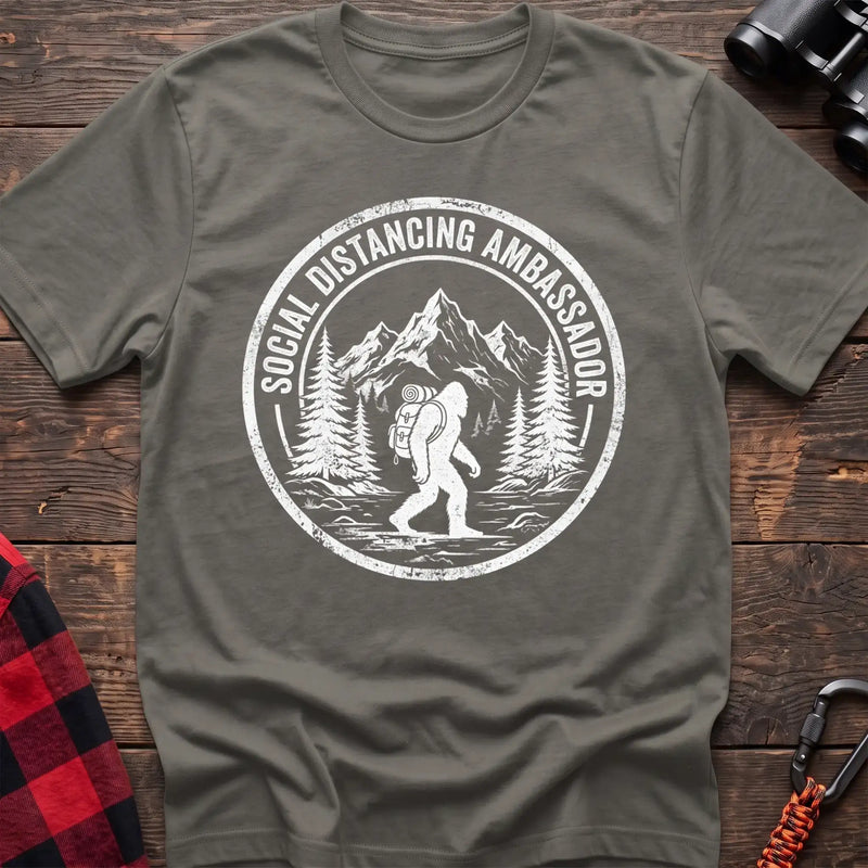 Heather military green t-shirt with white circular badge graphic of Bigfoot hiking with a backpack through mountains and the text Social Distancing Ambassador