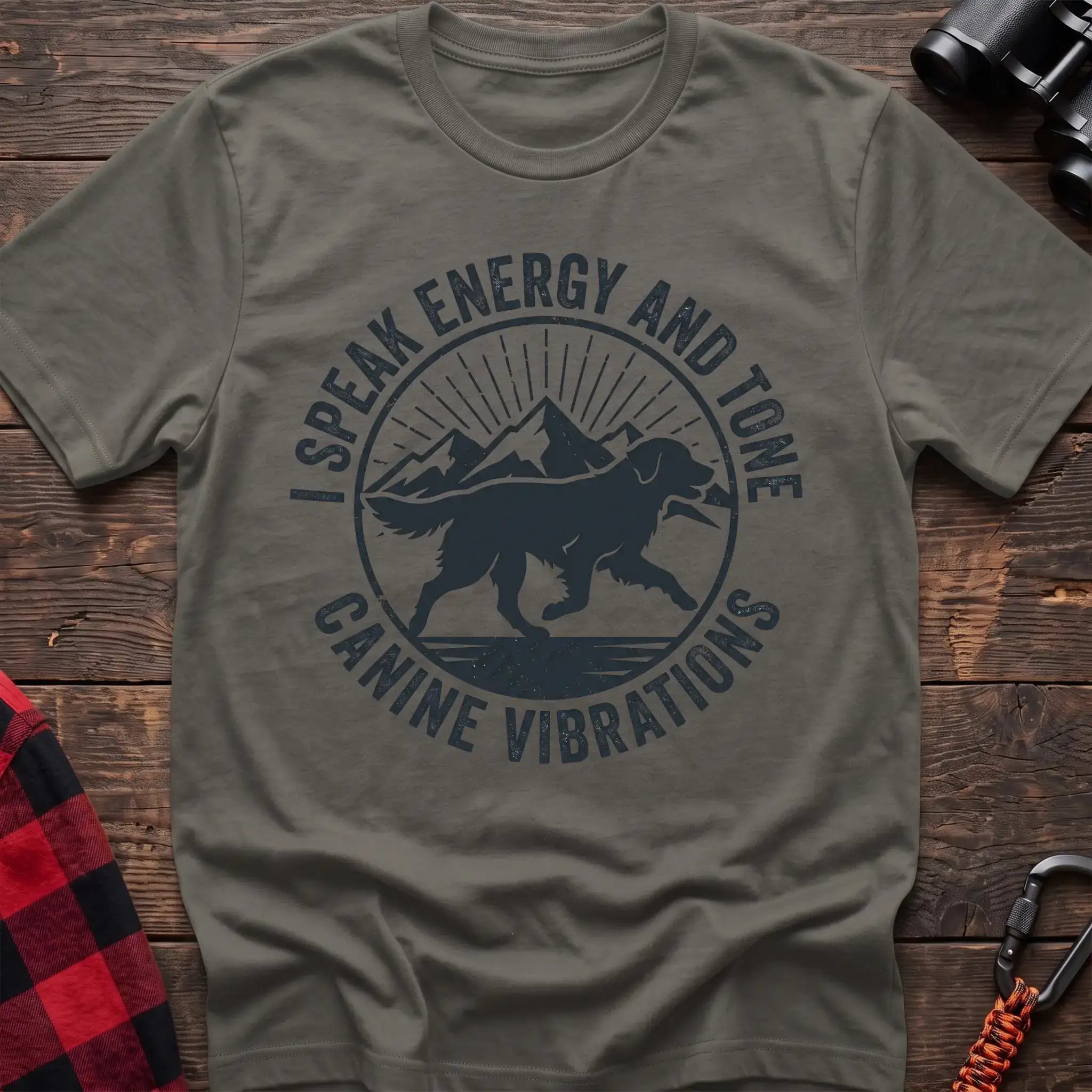 Heather military green t-shirt with a circular dark gray badge design featuring a running dog carrying a stick against mountain peaks and sun rays, with the text “I SPEAK ENERGY AND TONE” and “CANINE VIBRATIONS,” laid flat on a wooden surface.
