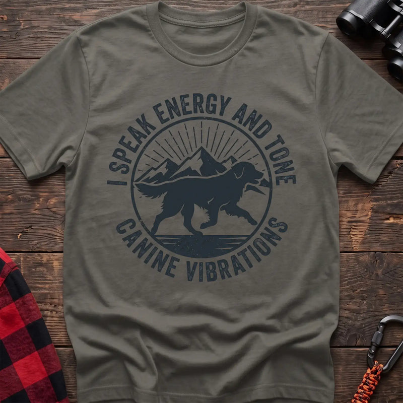Heather military green t-shirt with a circular dark gray badge design featuring a running dog carrying a stick against mountain peaks and sun rays, with the text “I SPEAK ENERGY AND TONE” and “CANINE VIBRATIONS,” laid flat on a wooden surface.