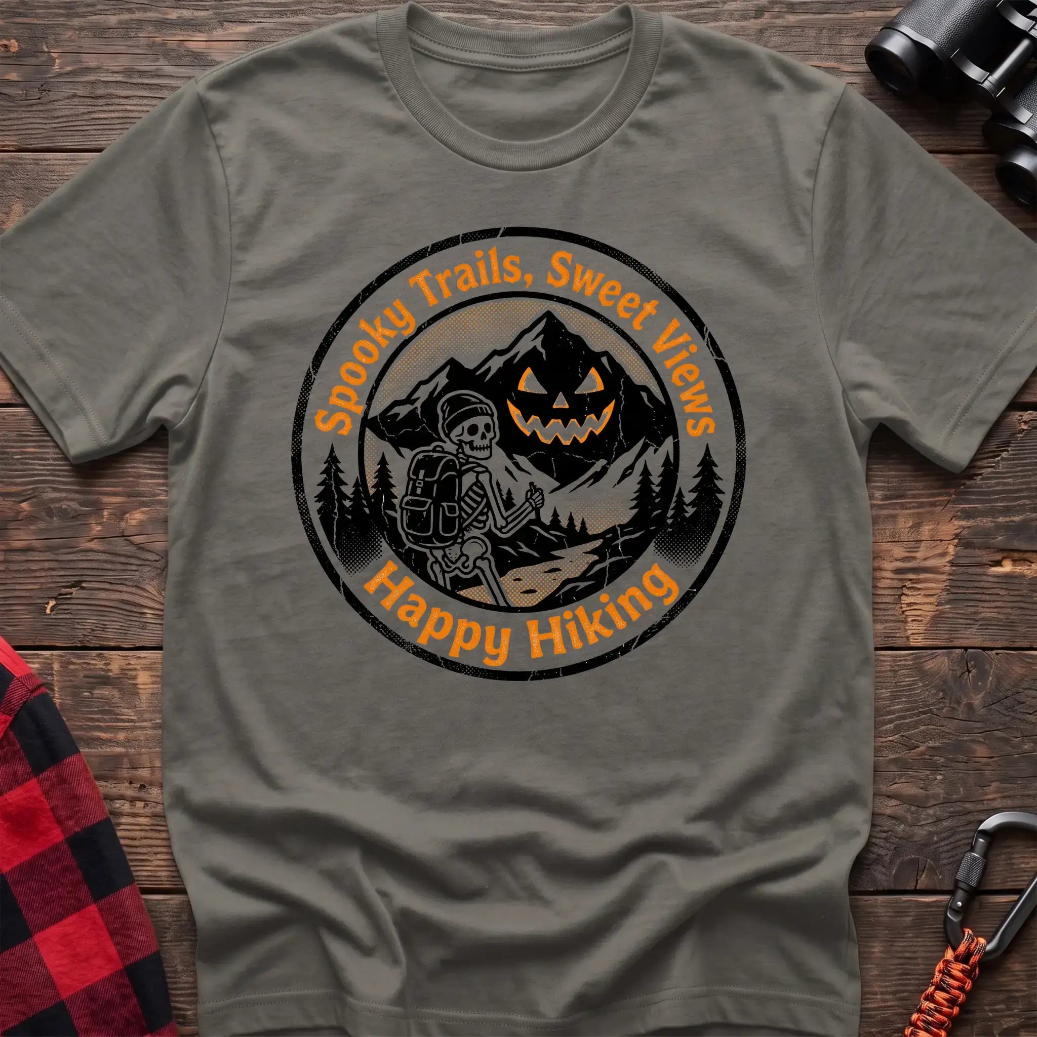 Heather military green t-shirt with circular graphic of a skeleton hiker giving a thumbs up in front of a jack-o-lantern mountain, reading Spooky Trails Sweet Views Happy Hiking