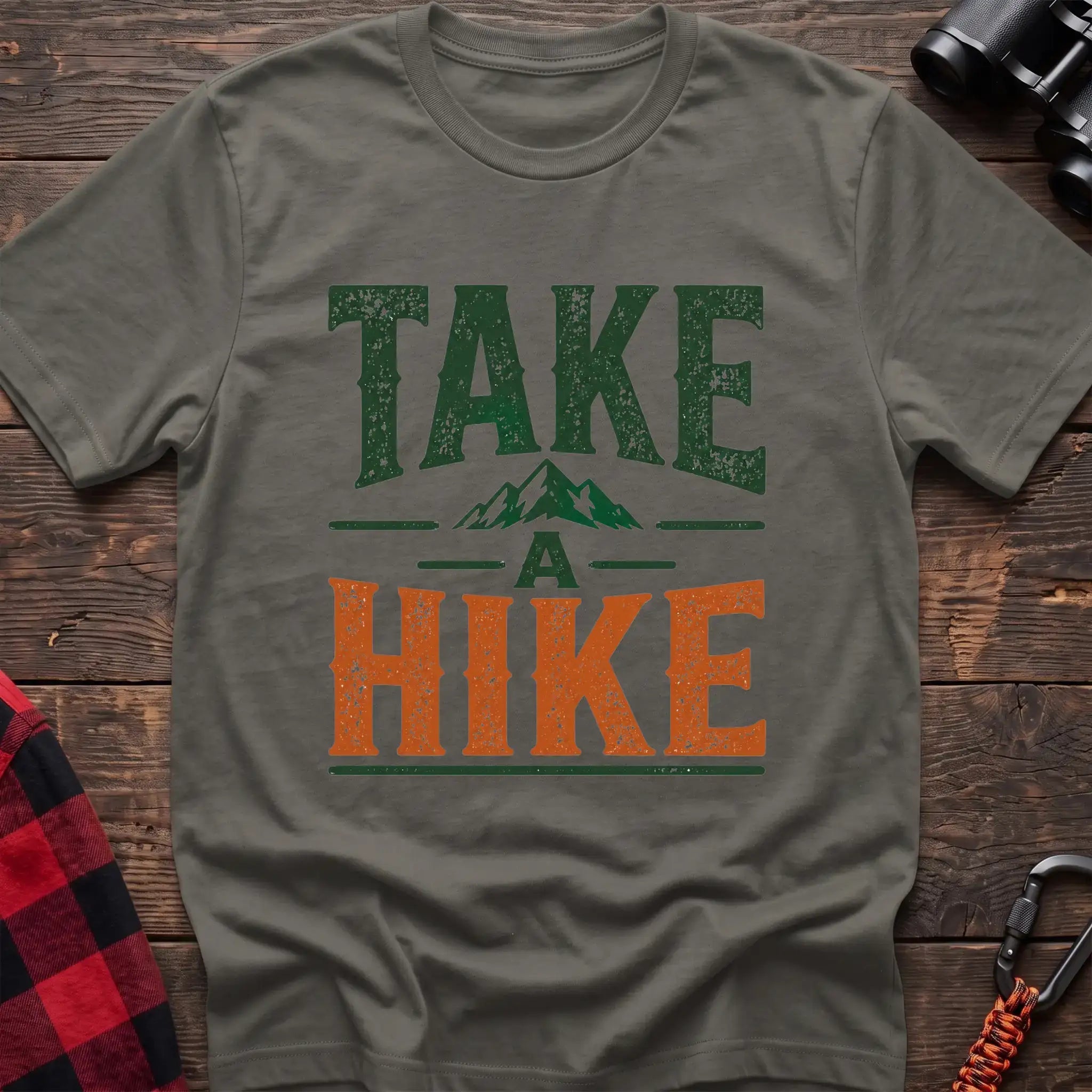 Heather military green t-shirt with distressed green and orange text reading “TAKE A HIKE” with a small mountain graphic between the words, laid flat on a wooden surface.