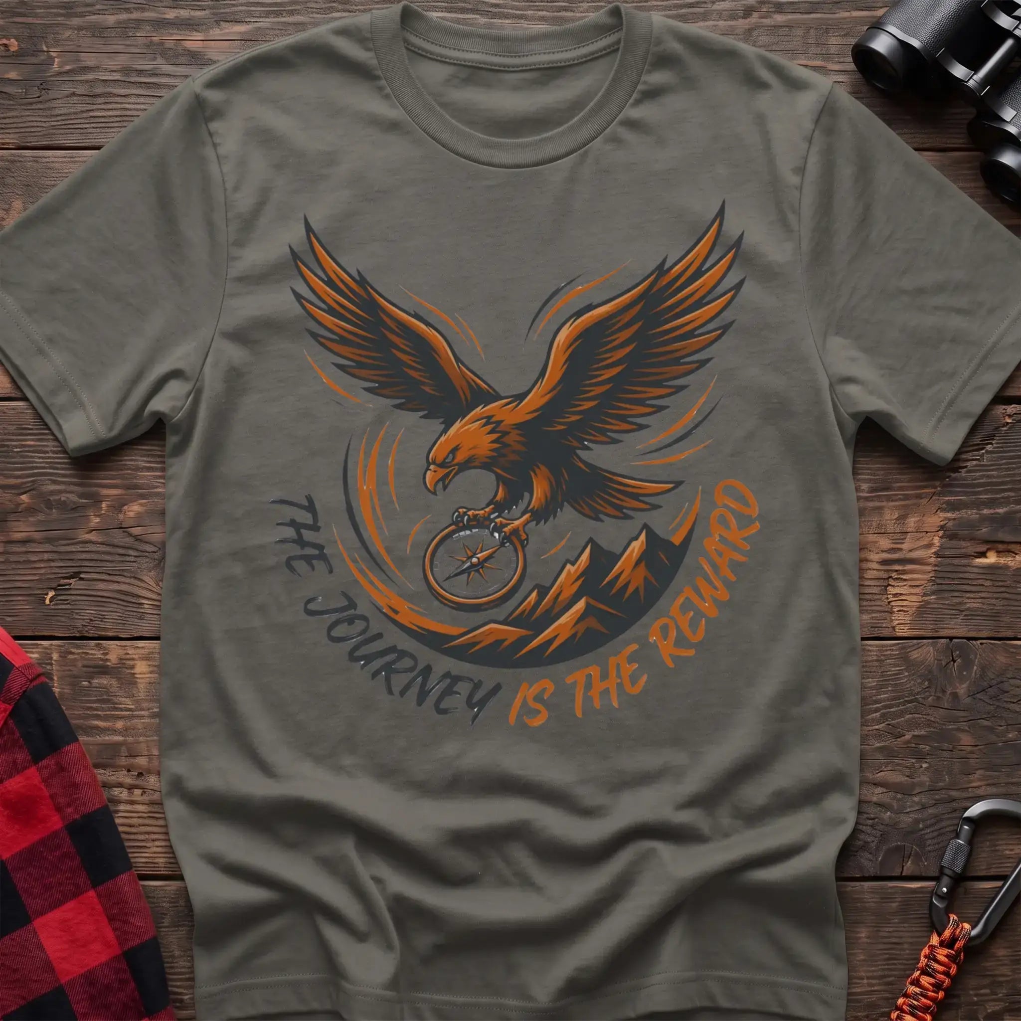 Heather military green t-shirt with an orange and dark graphic of a soaring eagle holding a compass above mountains, with the curved text “THE JOURNEY IS THE REWARD,” laid flat on a wooden surface.