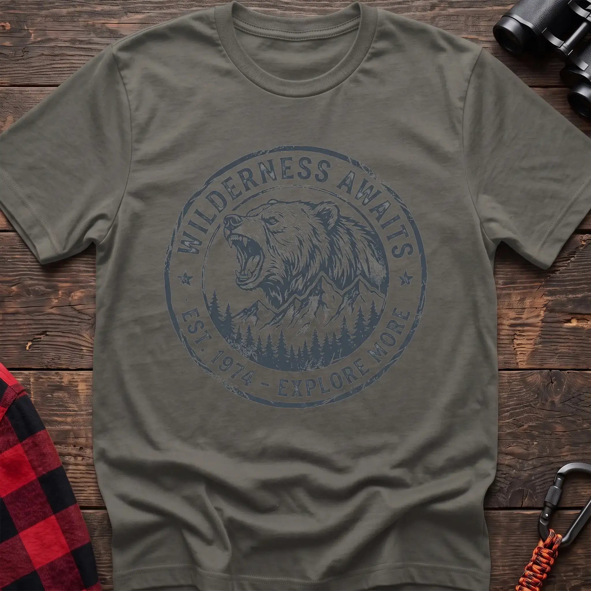 Heather military green t-shirt with a circular dark gray badge design featuring a roaring bear head above mountains and pine trees, with the text “WILDERNESS AWAITS” and “EST. 1974 - EXPLORE MORE,” laid flat on a wooden surface.