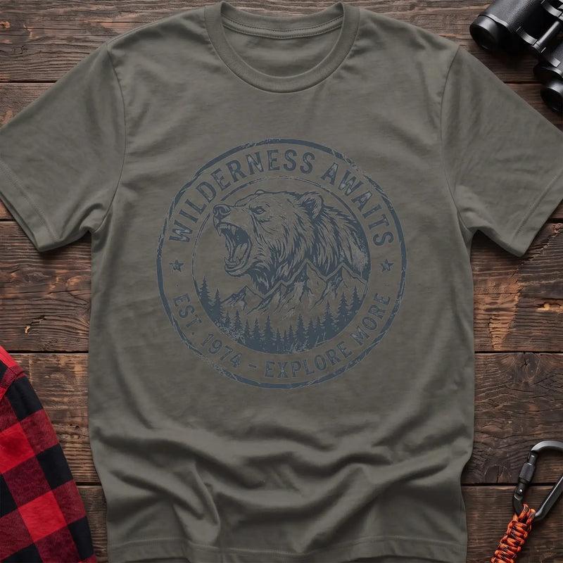 Heather military green t-shirt with a circular dark gray badge design featuring a roaring bear head above mountains and pine trees, with the text “WILDERNESS AWAITS” and “EST. 1974 - EXPLORE MORE,” laid flat on a wooden surface.