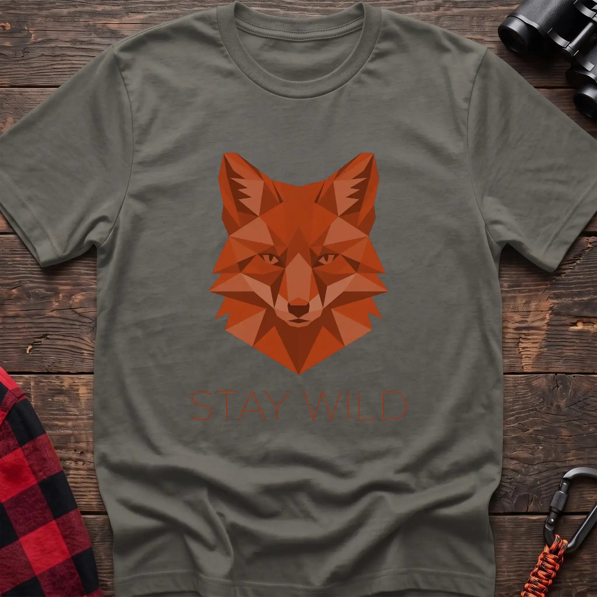 Heather military green t-shirt with a geometric low-poly orange fox head and the text “STAY WILD” printed below in matching orange, laid flat on a wooden surface.