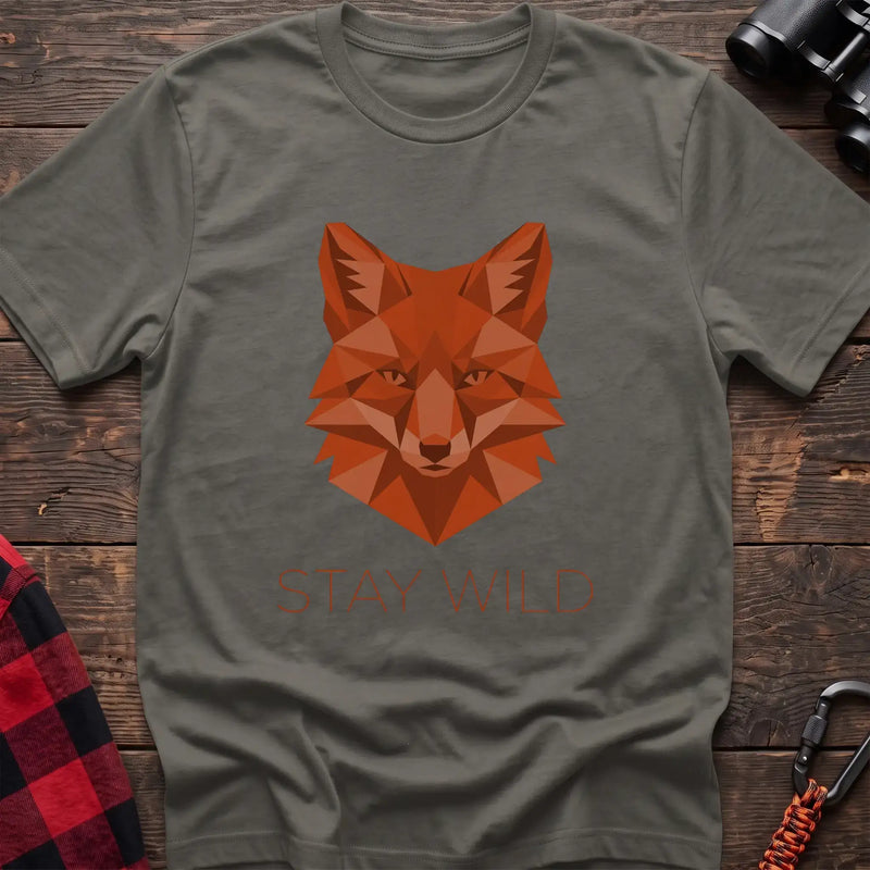 Heather military green t-shirt with a geometric low-poly orange fox head and the text “STAY WILD” printed below in matching orange, laid flat on a wooden surface.