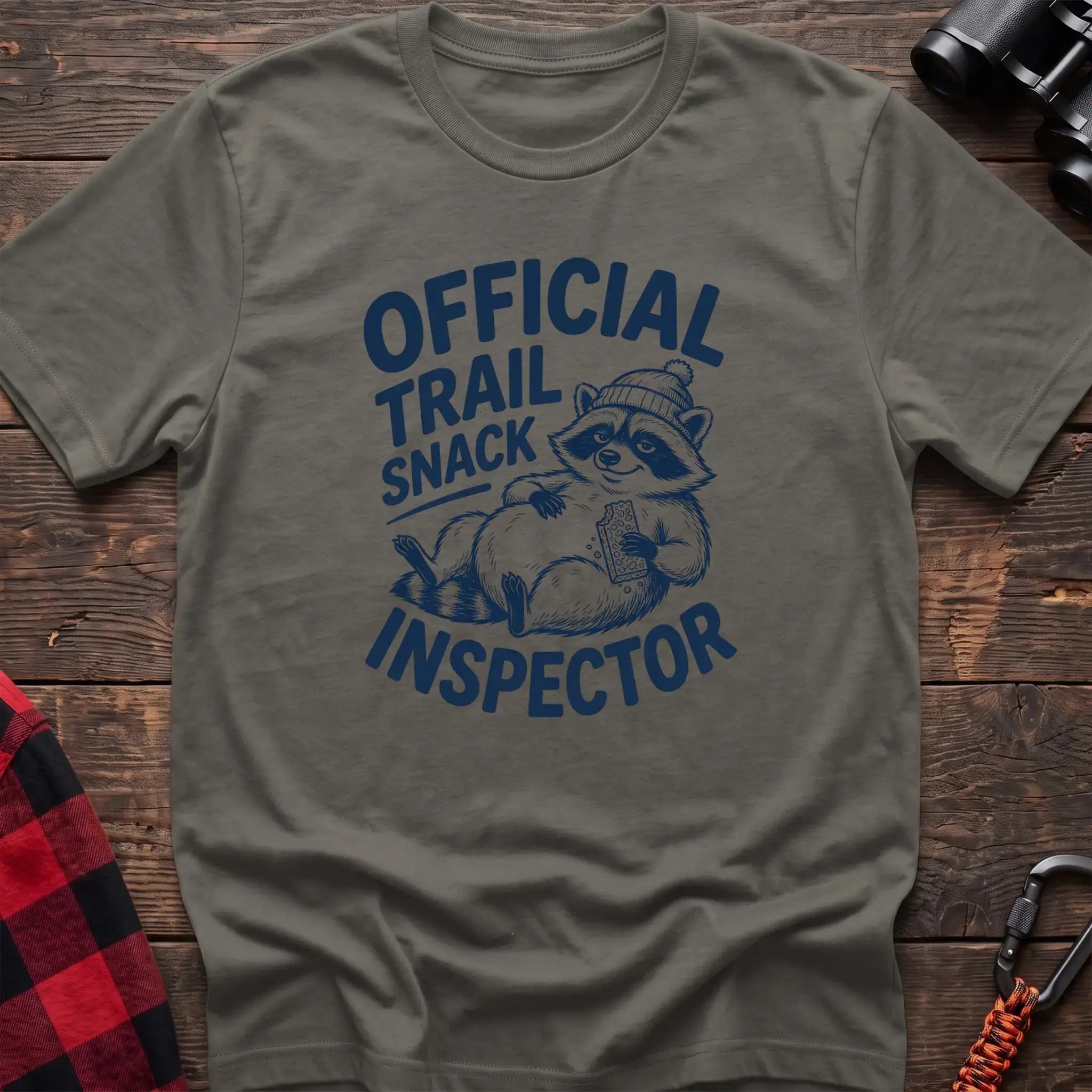 Heather military green t-shirt with cartoon raccoon holding a snack and the text Official Trail Snack Inspector