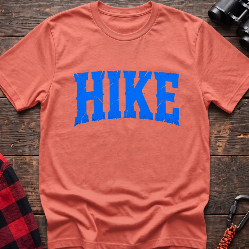Heather orange cotton t-shirt with distressed arched blue HIKE graphic in cracked varsity lettering, flat lay on rustic wood with hiking gear