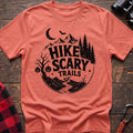 Heather orange cotton t-shirt with vintage Hike Scary Trails graphic of mountains, a crescent moon, bat, jack-o-lanterns, and a winding trail, flat lay on rustic wood with hiking gear